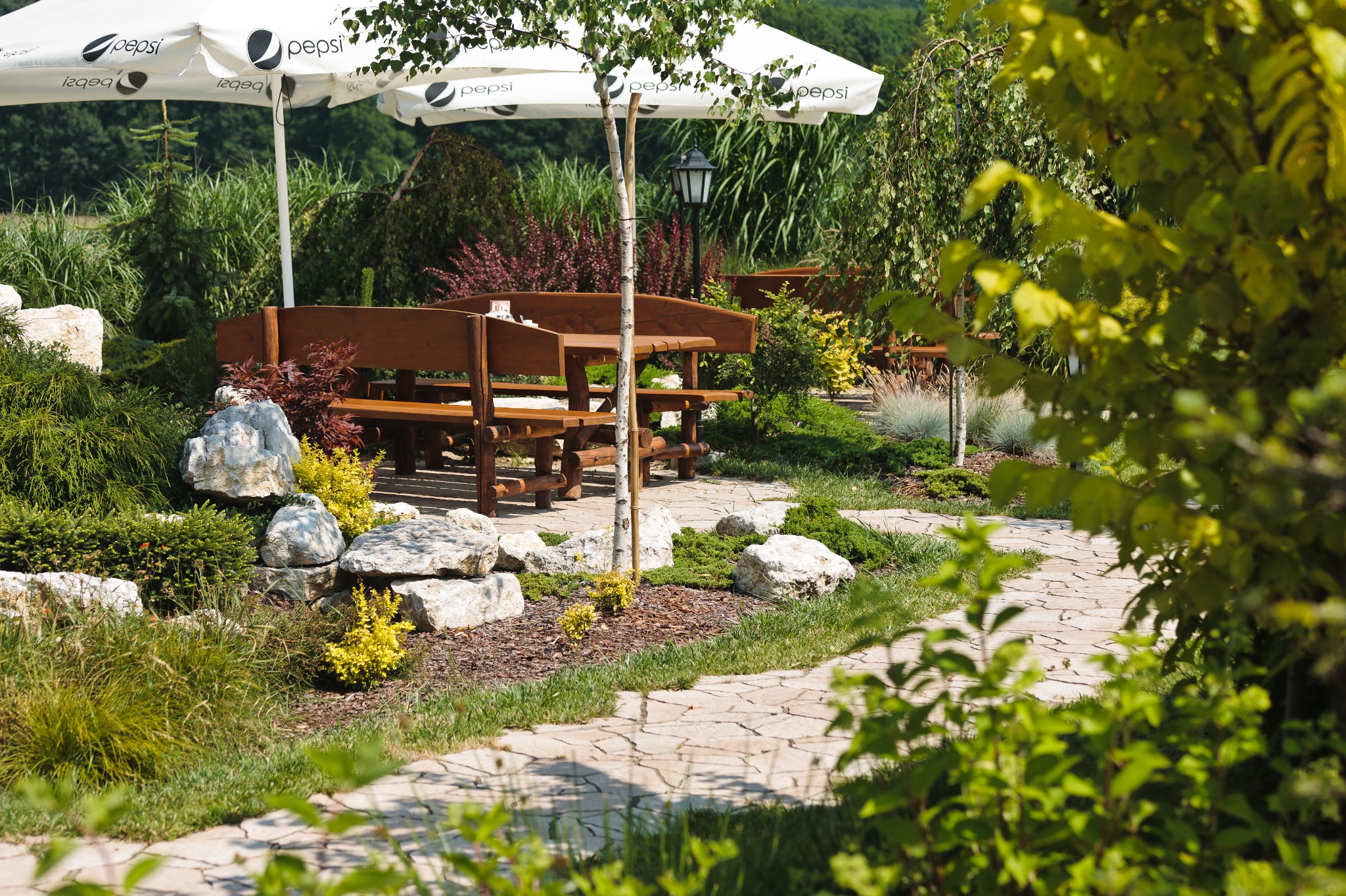 Urokliwe miejsce na zewnątrz z drewnianymi ławkami i stołami pod parasolami Pepsi, otoczone zielenią, kamieniami i ścieżką z płyt kamiennych, idealne na imprezę plenerową.