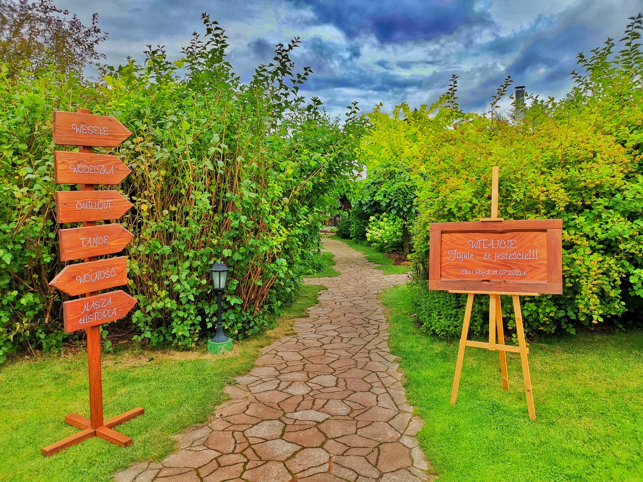 Ogród z kamienną ścieżką prowadzącą do tablicy powitalnej na sztaludze oraz drewnianego drogowskazu z napisem 'Wesele', 'Wódeczka', 'Chillout', 'Tańce', 'Wolność', 'Nasza Historia' na tle zieleni...
