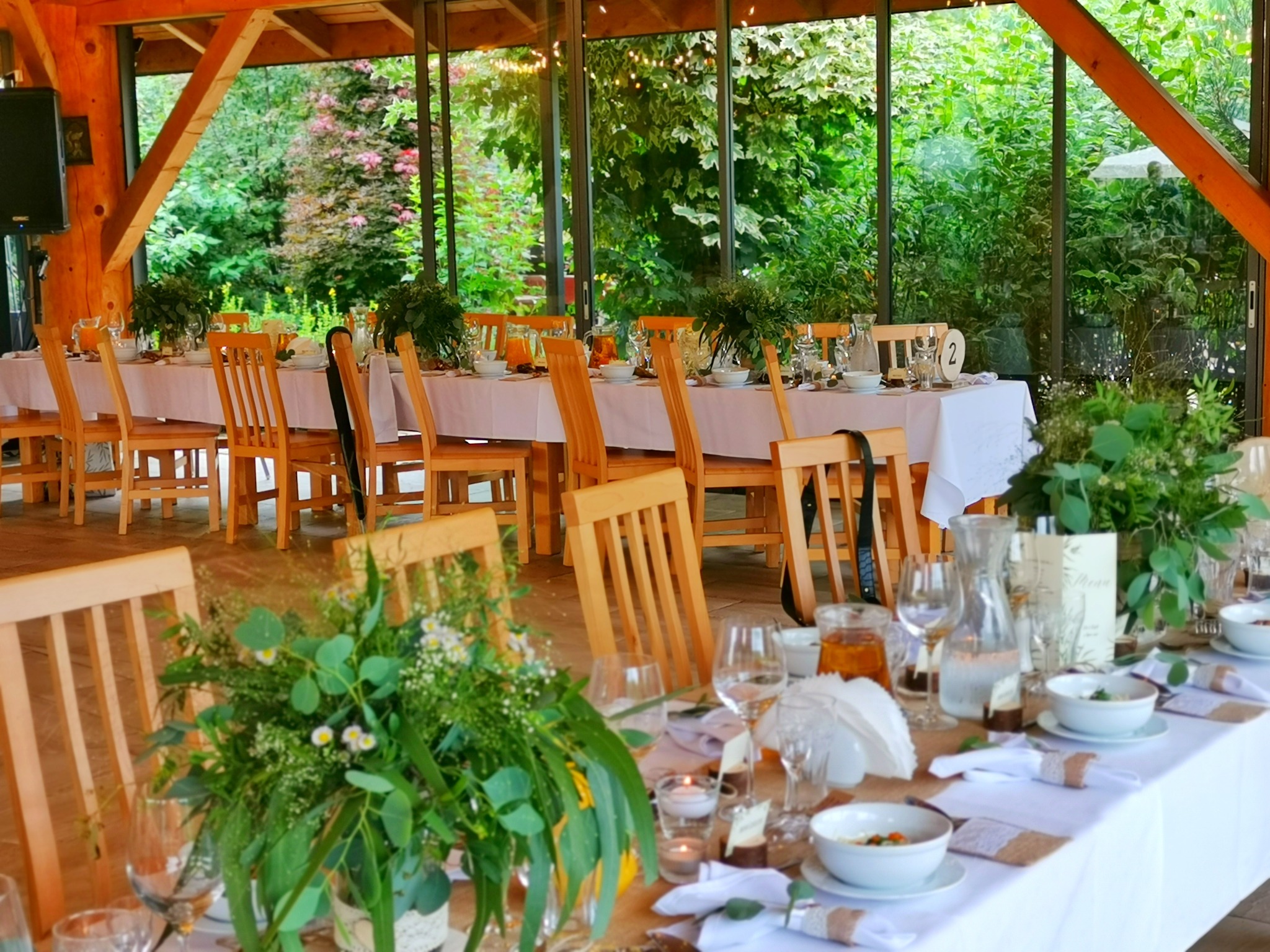 Uroczysta aranżacja stołu w letniej restauracji, z drewnianymi krzesłami i dekoracjami z naturalnych kwiatów i zieleni, widok na bujny ogród przez przeszkloną ścianę.