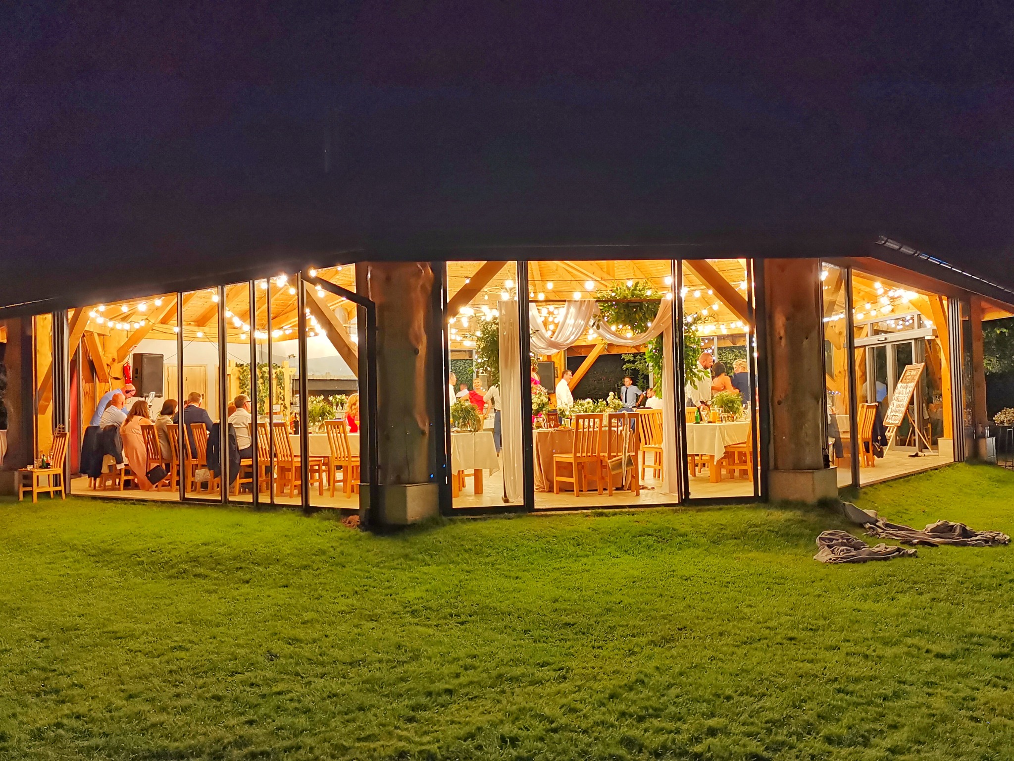 Oświetlona restauracja z dużymi oknami, widoczni goście siedzący przy stołach, dekoracje roślinne i girlandy świetlne, trawnik przed budynkiem, wieczorny krajobraz.