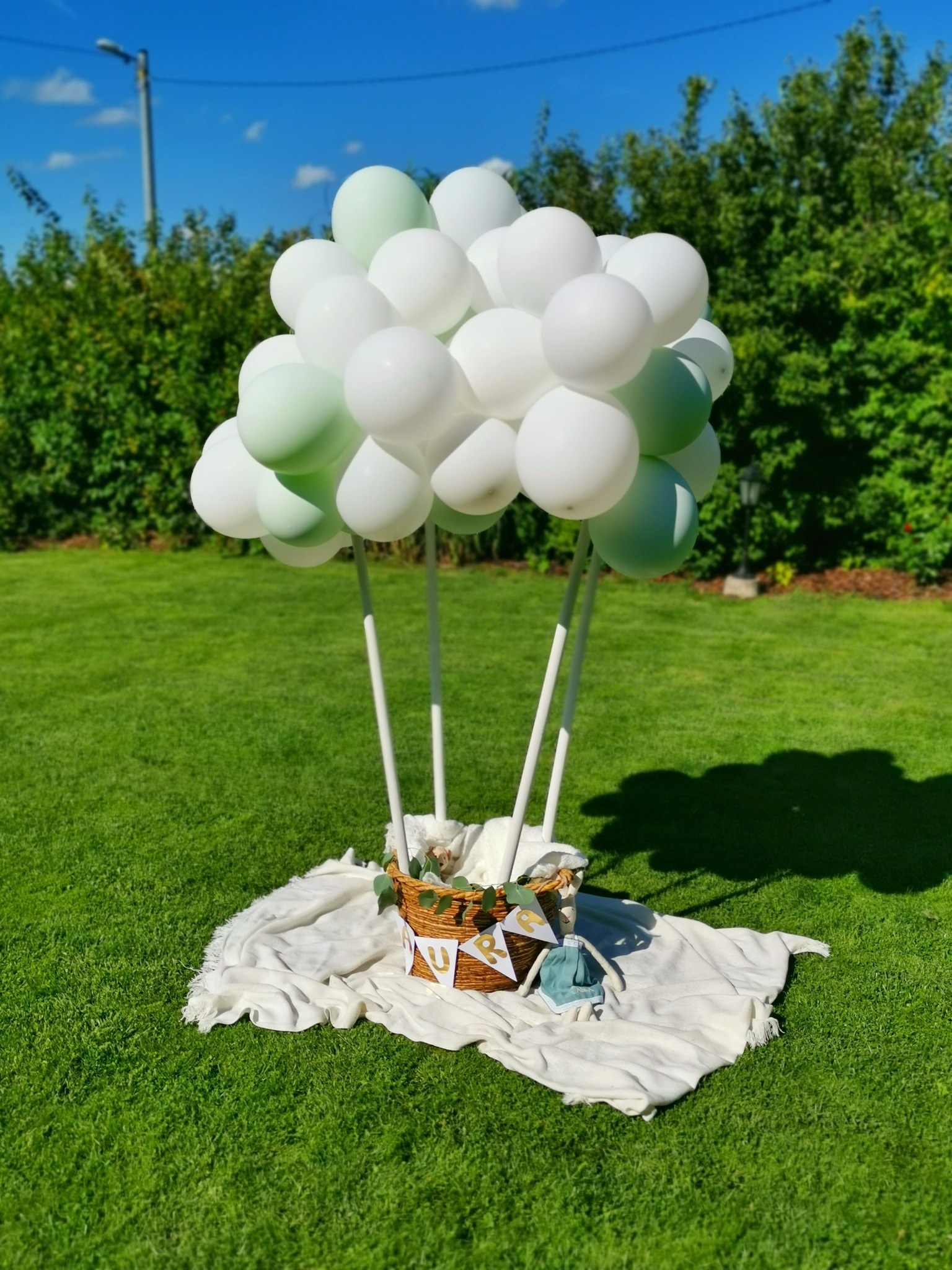 Dekoracja balonowa w kształcie balonu na ogród. Białe i jasnozielone balony, kosz wiklinowy z dekoracją i pluszową zabawką na białym kocu, na tle zielonego trawnika i żywopłotu.