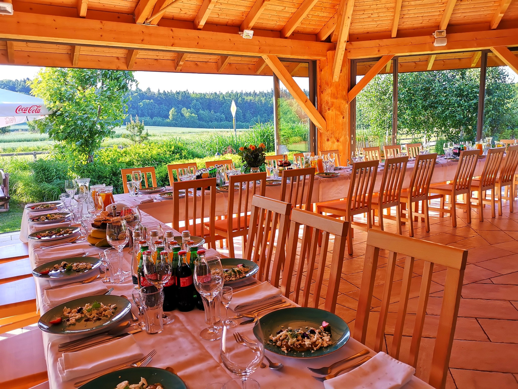 Długi stół nakryty do przyjęcia w drewnianej altanie z widokiem na zielone pola i las, z napojami Coca-Cola i talerzami z sałatką.
