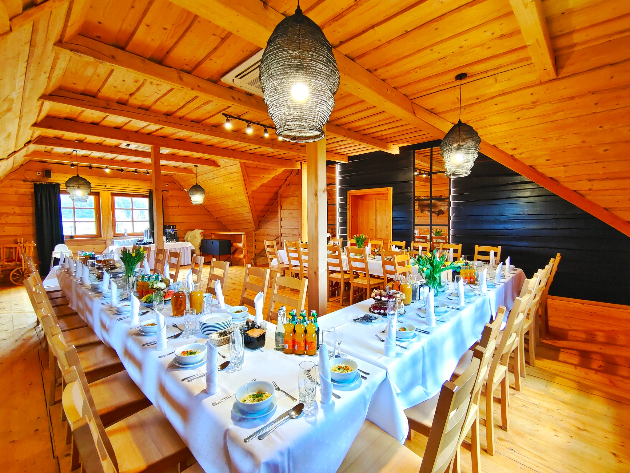 Uroczysta sala w stylu rustykalnym z drewnianym stropem i długimi stołami nakrytymi białymi obrusami, gotowymi na przyjęcie gości, oświetlona plecionymi lampami wiszącymi.