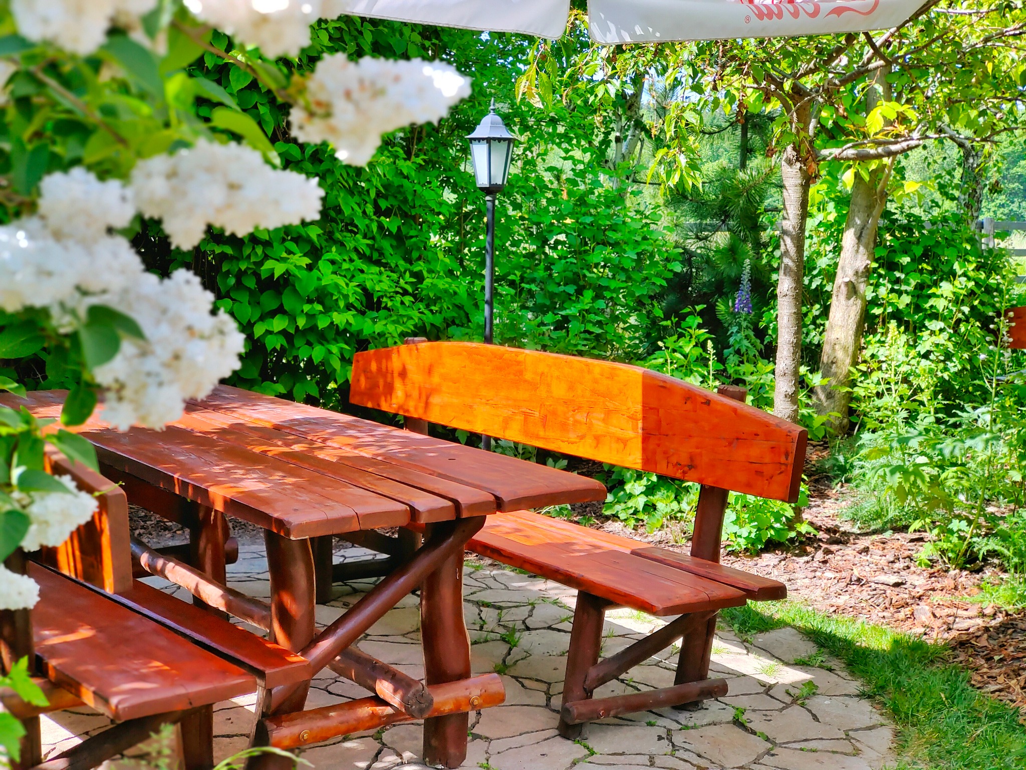 Rustykalny drewniany stół z ławkami w ogrodzie, oświetlony latarnią, z kwitnącym bzem w tle, na kamiennej ścieżce.