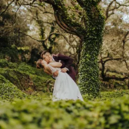 Pocałunek pary młodej w lesie, panna młoda w koronkowej sukni, pan młody w bordowym garniturze, drzewo porośnięte bluszczem w tle.