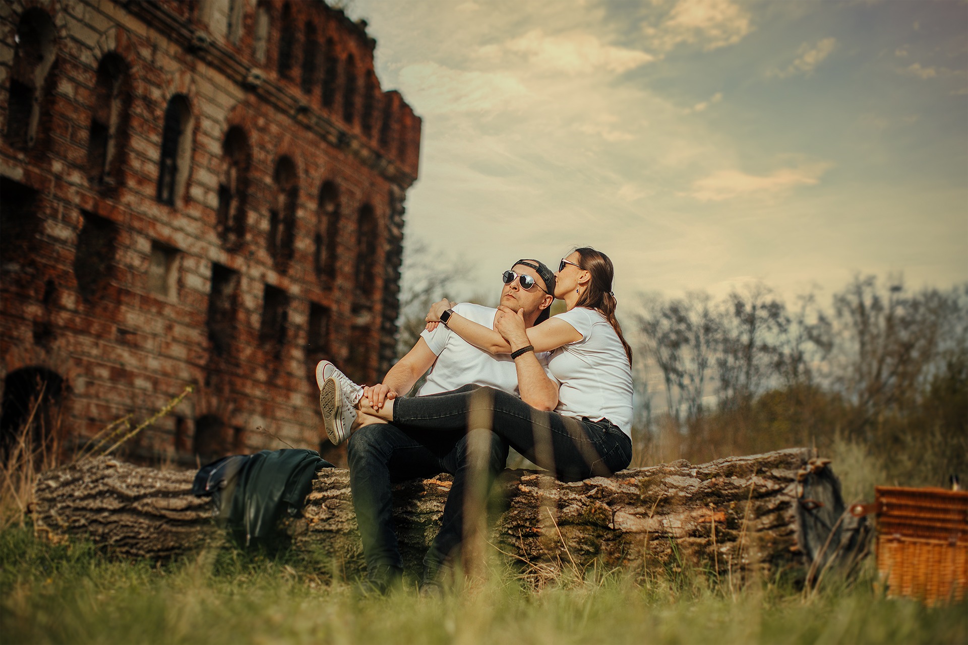 Para w okularach przeciwsłonecznych siedzi na powalonym pniu drzewa na tle starego, ceglanego budynku z łukowymi oknami, w oddali widoczne drzewa i zachmurzone niebo.