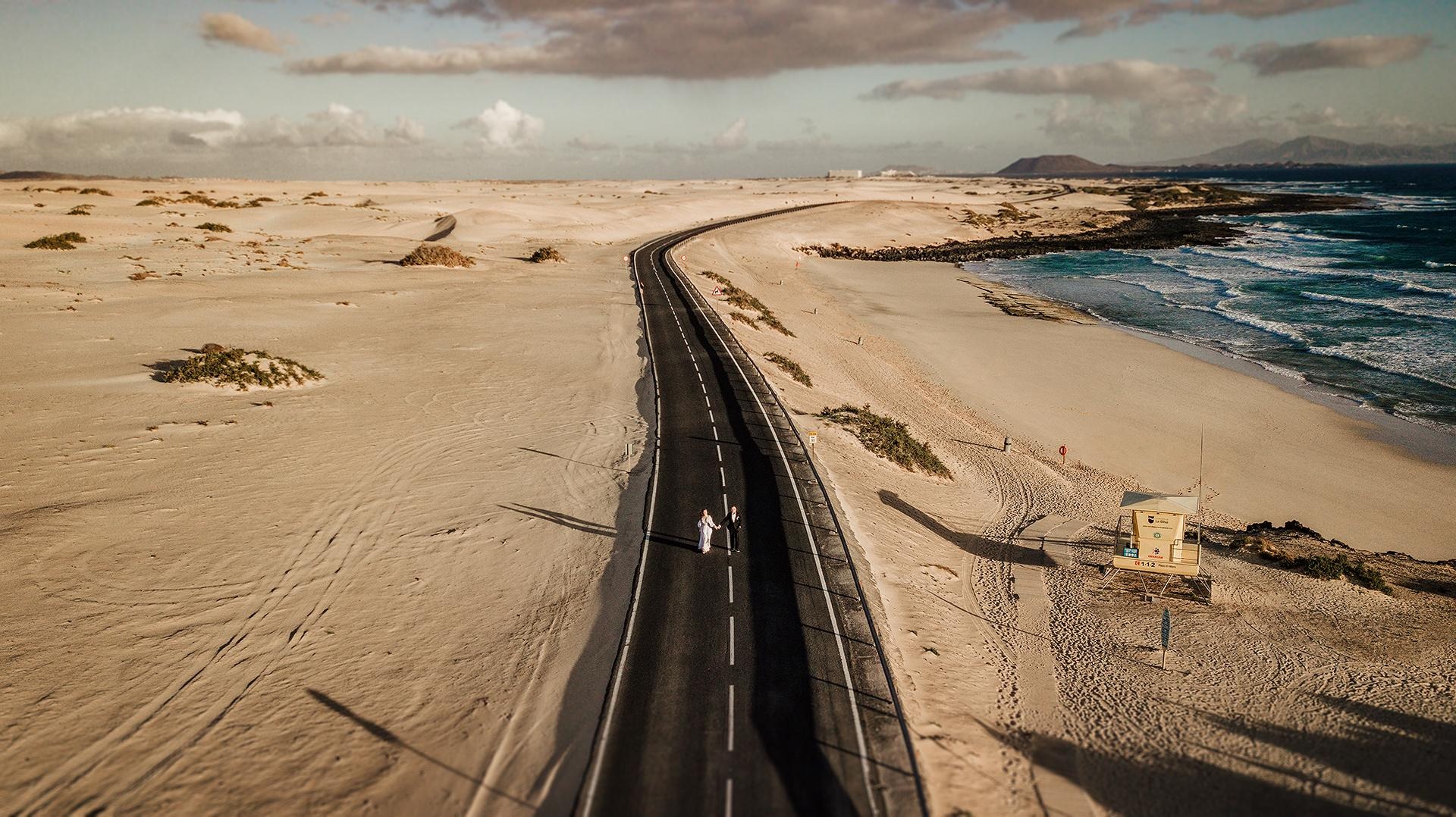 Zdjęcie z lotu ptaka przedstawia parę młodą idącą po czarnej drodze przecinającej piaszczystą pustynię w kierunku morza z wieżą ratowniczą na plaży.