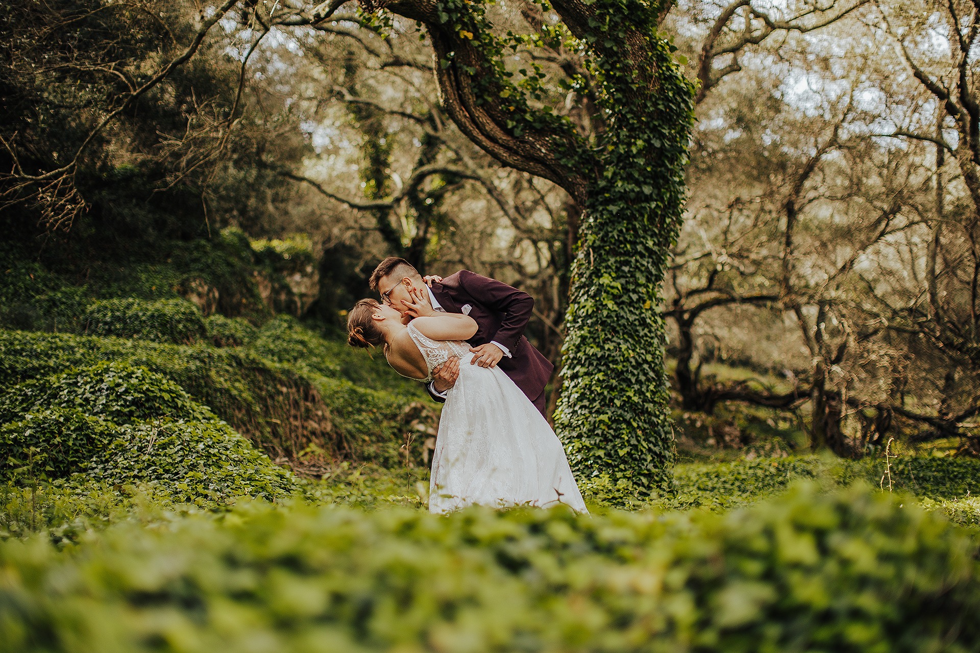 Pocałunek pary młodej w lesie, panna młoda w koronkowej sukni, pan młody w bordowym garniturze, drzewo porośnięte bluszczem w tle.