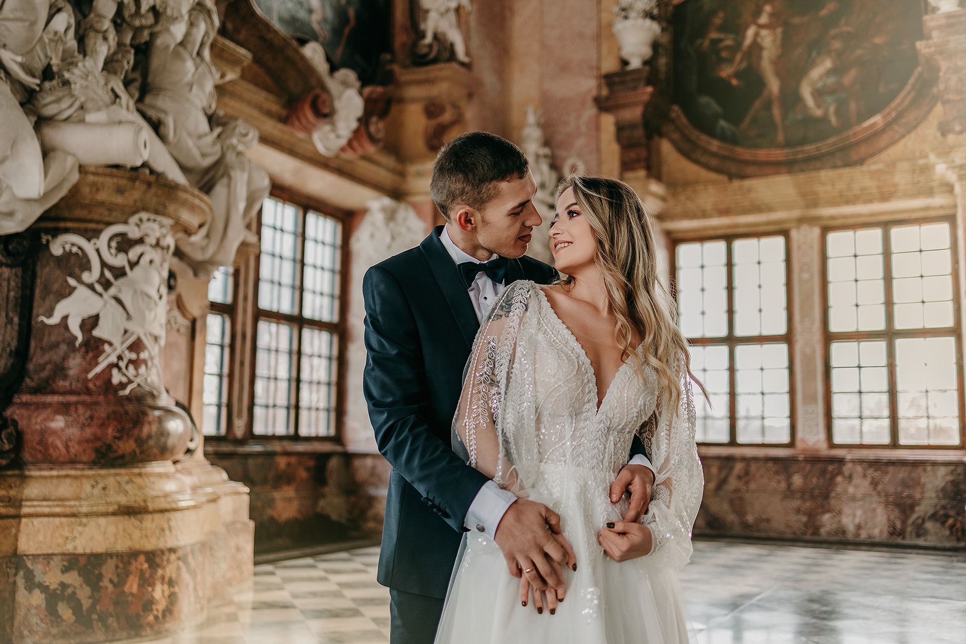 Para młoda w objęciach na tle barokowego wnętrza z marmurowymi kolumnami i obrazami, panna młoda w sukni z koronkowymi rękawami i głębokim dekoltem, pan młody w granatowym smokingu z muchą.