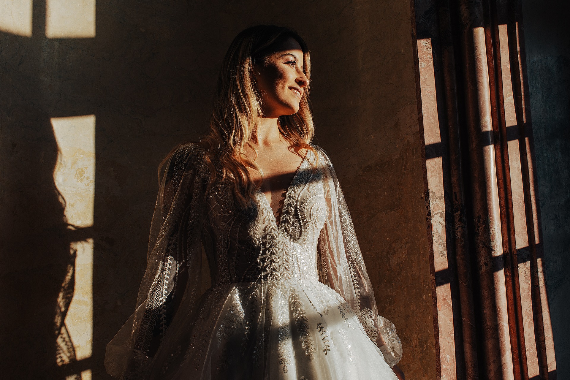 Uśmiechnięta panna młoda w koronkowej sukni z długimi rękawami, oświetlona promieniami słońca wpadającymi przez okno z marmurowymi ramami.