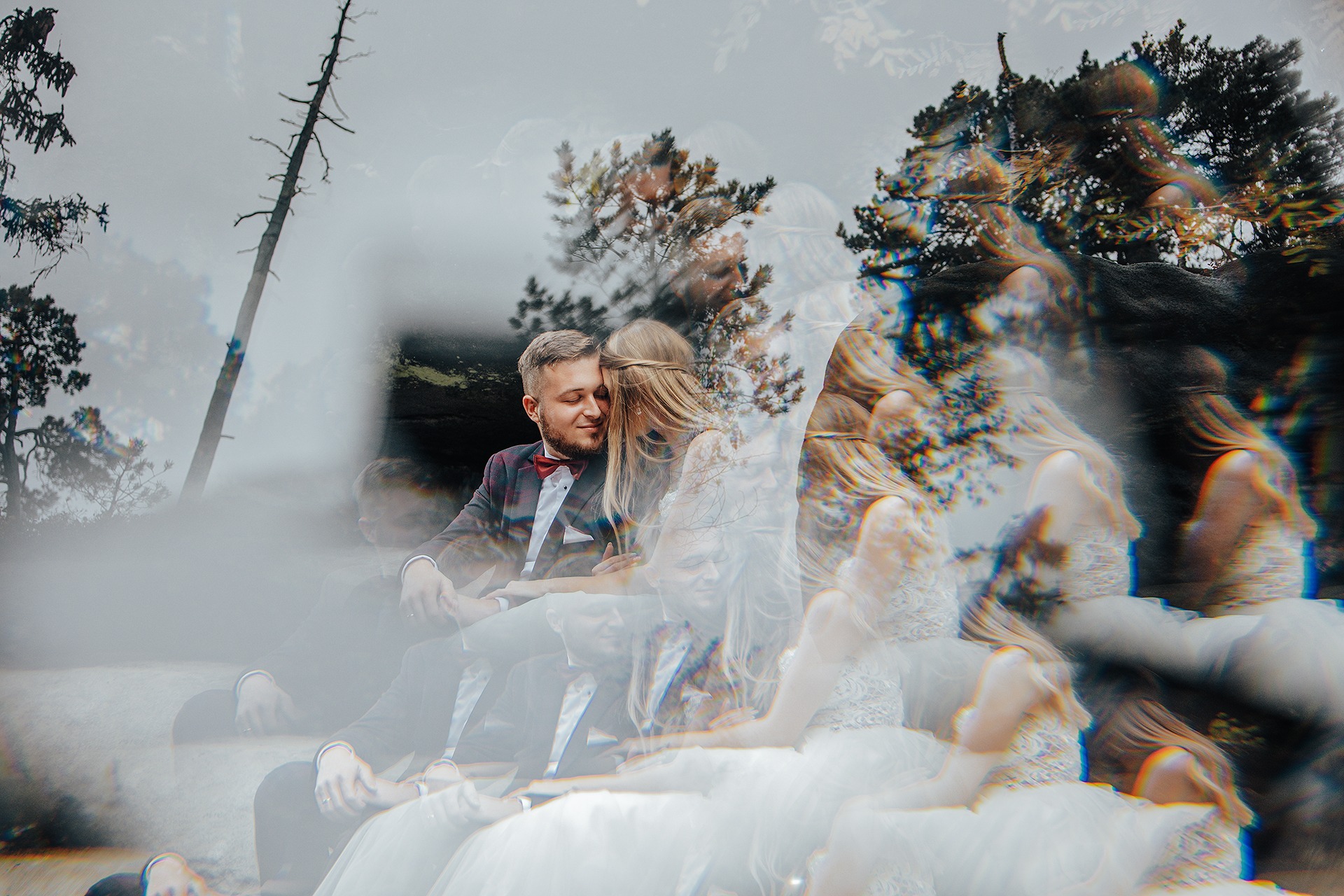 Młoda para w strojach ślubnych siedzi blisko siebie na tle drzew i pochmurnego nieba, z efektem nałożonych warstw i rozmycia, tworzącym artystyczny, eteryczny portret.