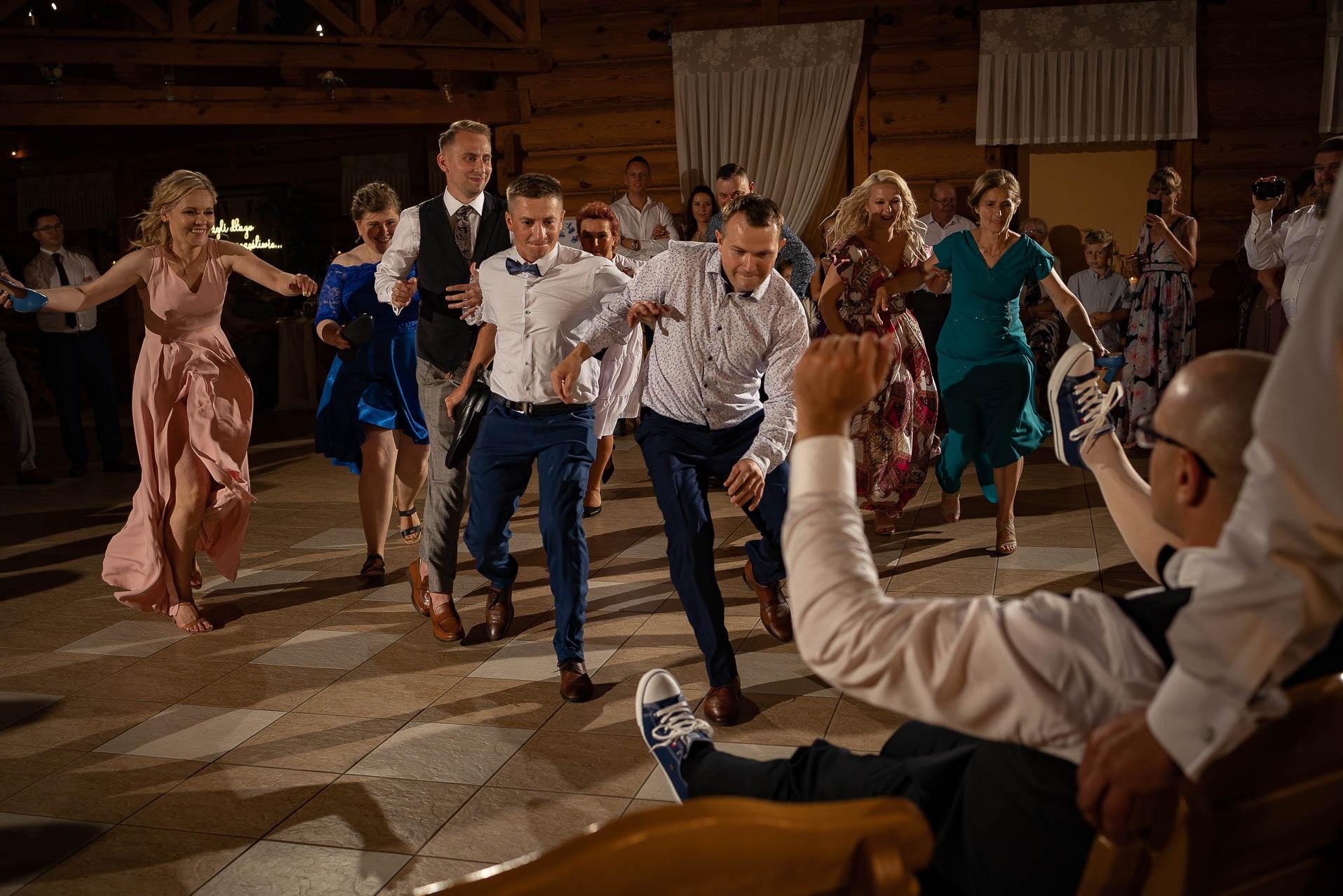 Radosna zabawa taneczna gości weselnych w Pajęcznie, uchwycona z perspektywy siedzącego mężczyzny trzymającego w górze niebieskiego trampka.