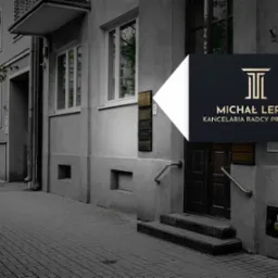 Zewnętrzny widok na wejście do kancelarii z czarnym szyldem z złotym logo i napisem Michał Lerka Kancelaria Radcy Prawnego, budynek w odcieniach szarości.
