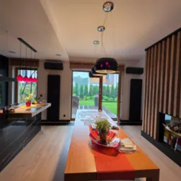 Nowoczesne wnętrze domu z klimatyzacją: czarne jednostki ścienne, minimalistyczne oświetlenie nad drewnianym stołem i widok na zadbany ogród przez przeszklone drzwi.