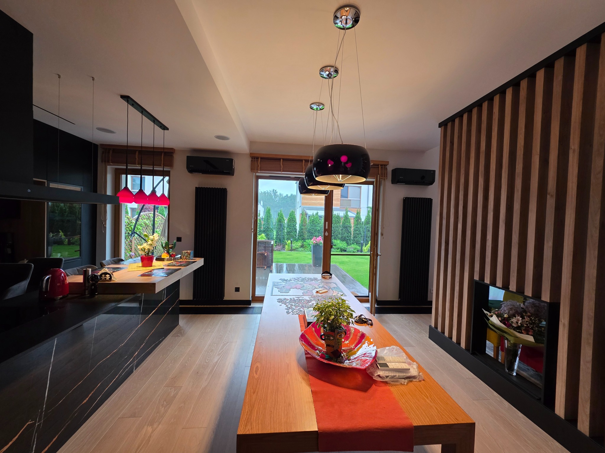 Nowoczesne wnętrze domu z klimatyzacją: czarne jednostki ścienne, minimalistyczne oświetlenie nad drewnianym stołem i widok na zadbany ogród przez przeszklone drzwi.