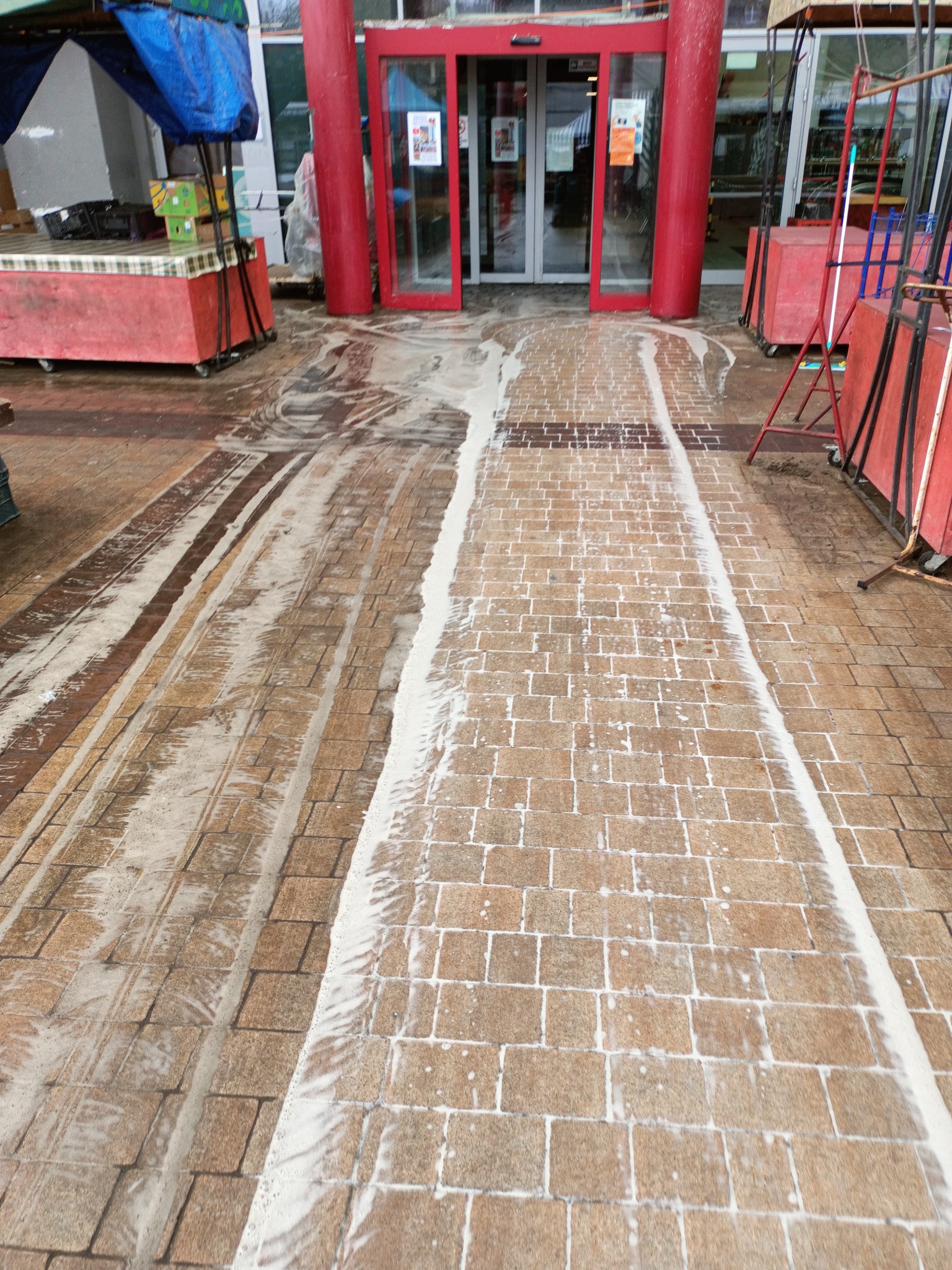 Częściowo umyta kostka brukowa przed wejściem do budynku z czerwonymi kolumnami i drzwiami, widoczne ślady detergentu i różnica w kolorze między czystą a brudną powierzchnią.