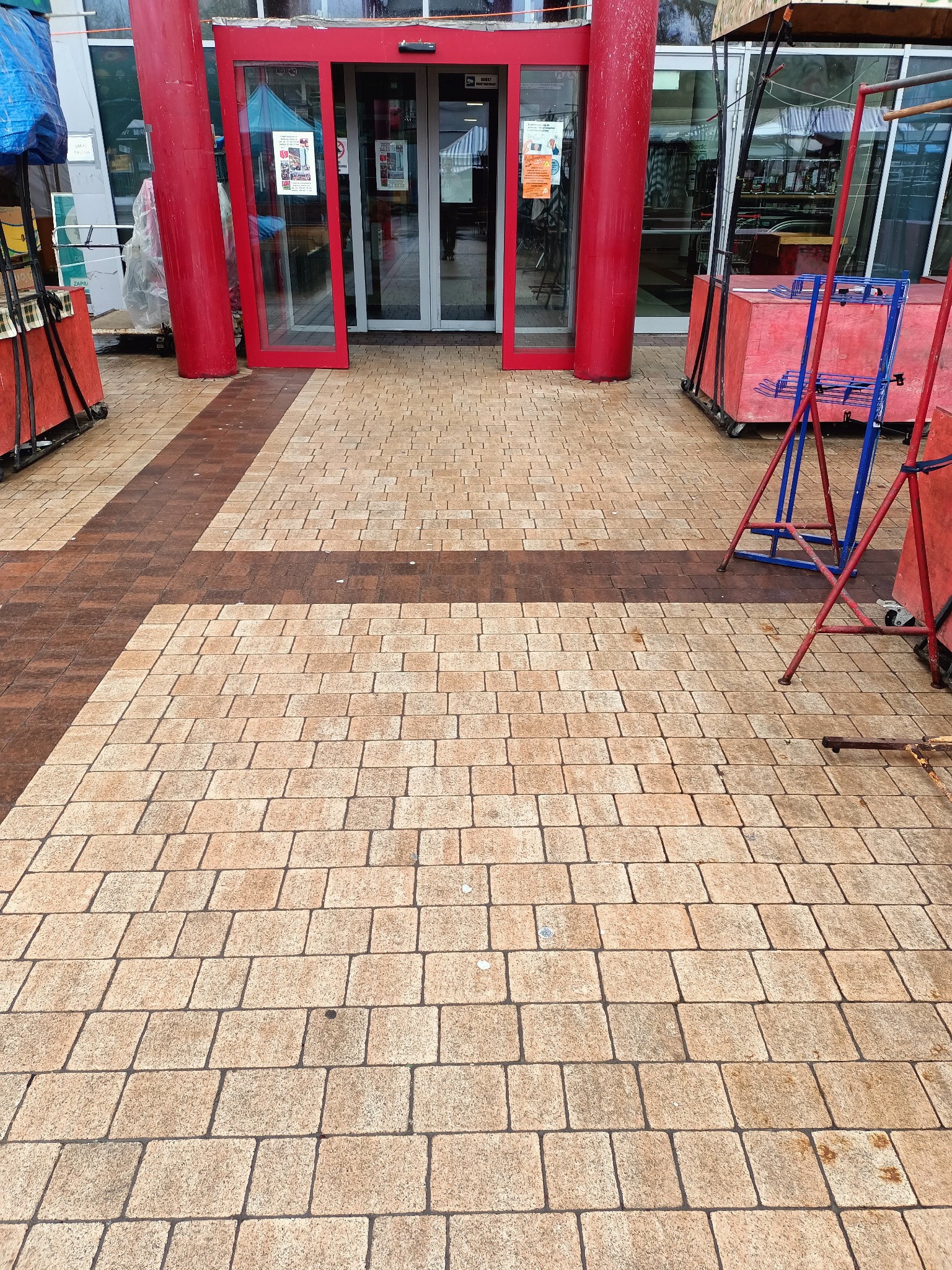 Ułożona kostka brukowa w dwóch odcieniach, tworząca wzór krzyża, prowadząca do wejścia z czerwonymi drzwiami i kolumnami, otoczonego przez tymczasowe konstrukcje handlowe.