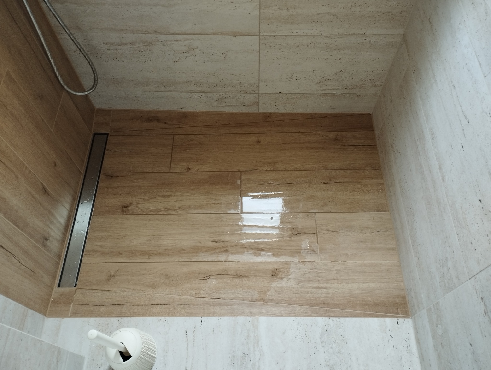 Wykończona kabina prysznicowa z odpływem liniowym, wyłożona płytkami imitującymi drewno na podłodze i jasnymi, beżowymi płytkami na ścianach, widoczny fragment słuchawki prysznicowej i szczotki...