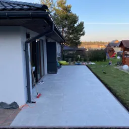 Nowoczesny taras z jasnoszarych płyt, obrzeża z ciemniejszej kostki, widok na ogród z elementami placu zabaw i dom w tle, wczesny wieczór.