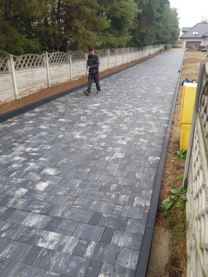 Długa, szara ścieżka z kostki brukowej prowadząca wzdłuż betonowego ogrodzenia, z pracownikiem idącym w oddali i żółtą skrzynką gazową przy ogrodzeniu.