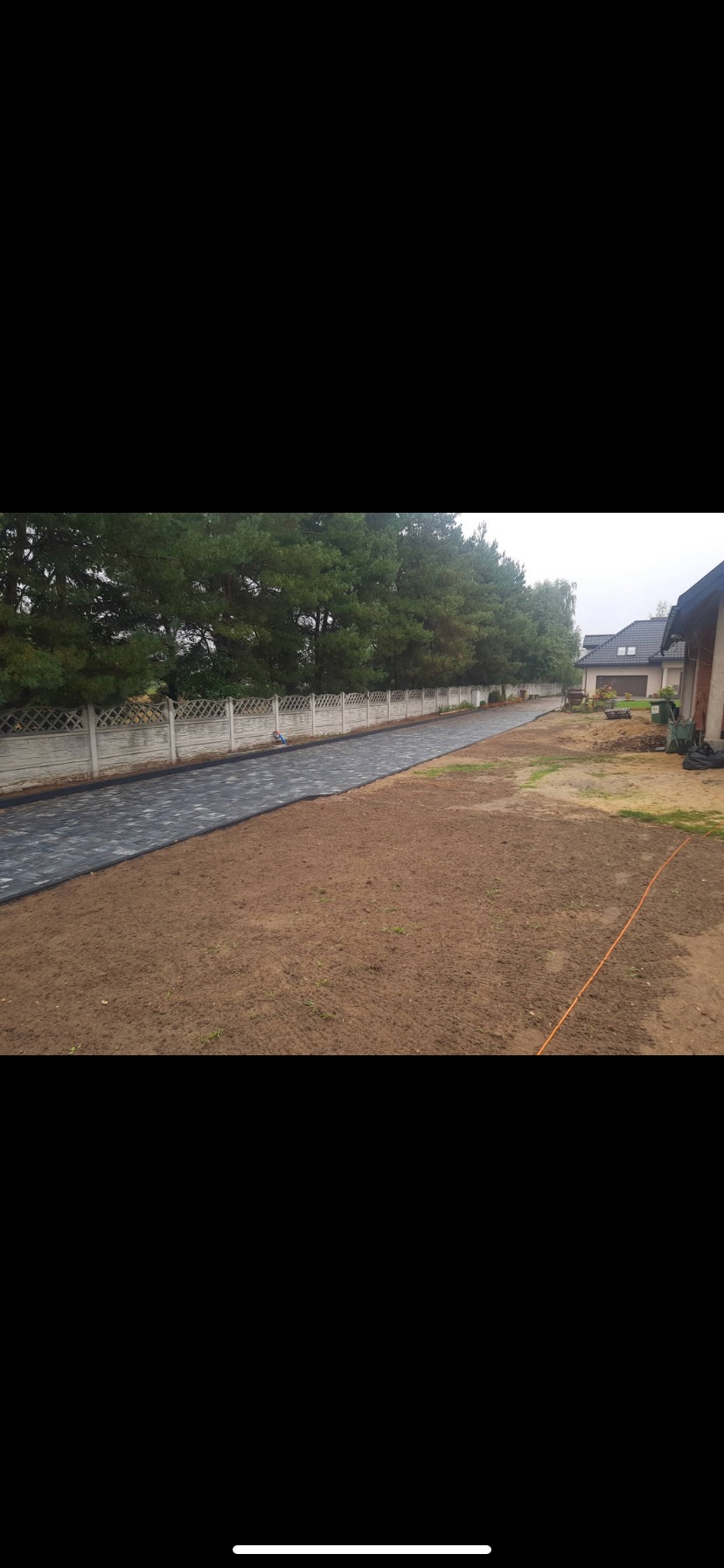 Częściowo ułożona, długa, ciemna kostka brukowa tworząca ścieżkę wzdłuż ogrodzenia z betonowych przęseł, na tle drzew i domu.