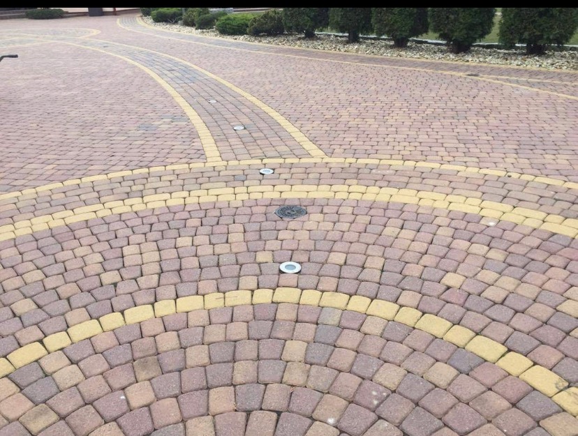 Ułożona kostka brukowa w odcieniach brązu i żółci tworząca wzór łuku i prostej ścieżki z wbudowanymi lampami oświetleniowymi, otoczona zielenią.