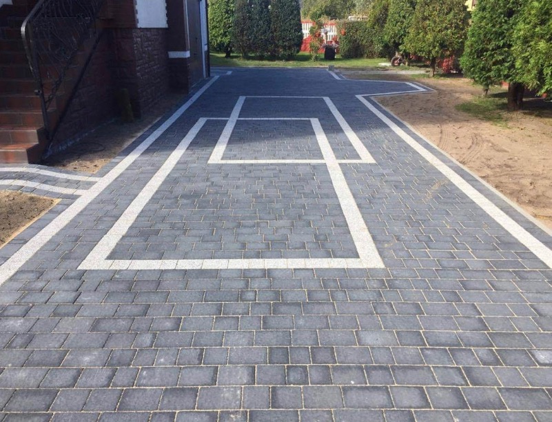Ułożona szara kostka brukowa z białymi obramowaniami tworzącymi geometryczny wzór przed domem, widoczne schody wejściowe i fragment ogrodu.