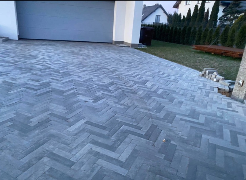 Ułożona szara kostka brukowa w jodełkę przed garażem, z zapasem kostki po prawej stronie i widocznym trawnikiem z tujami w tle.