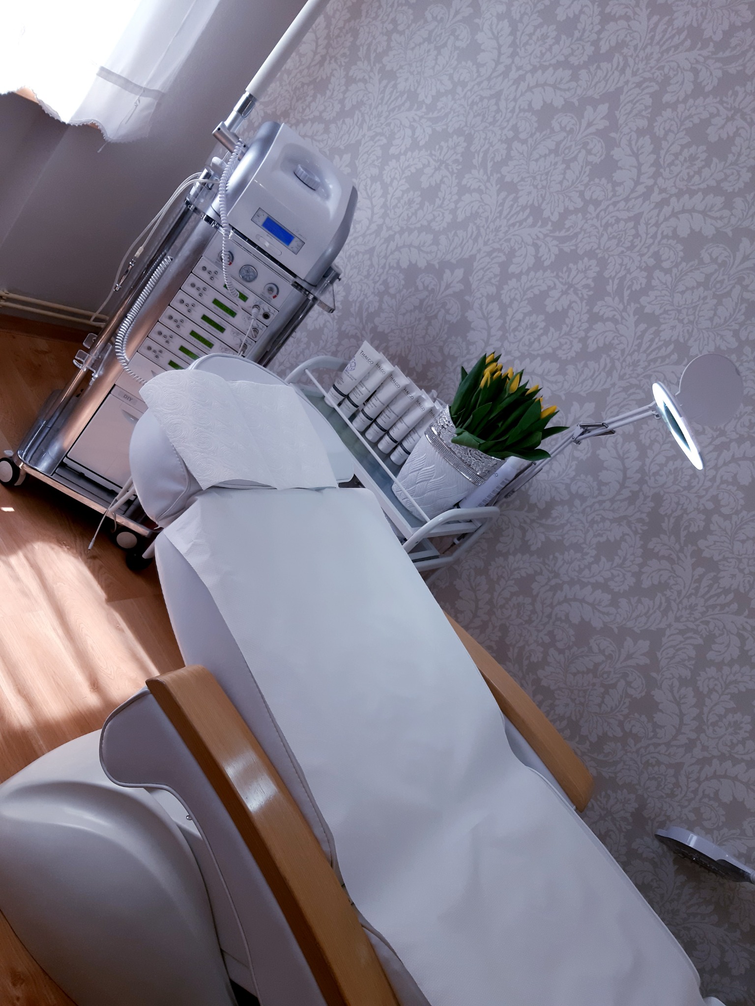 Przygotowane stanowisko do zabiegów kosmetycznych w Cieszynie: biały fotel z drewnianymi podłokietnikami przykryty jednorazowym prześcieradłem, obok wózek z aparaturą i kosmetykami Thalgo, a także...