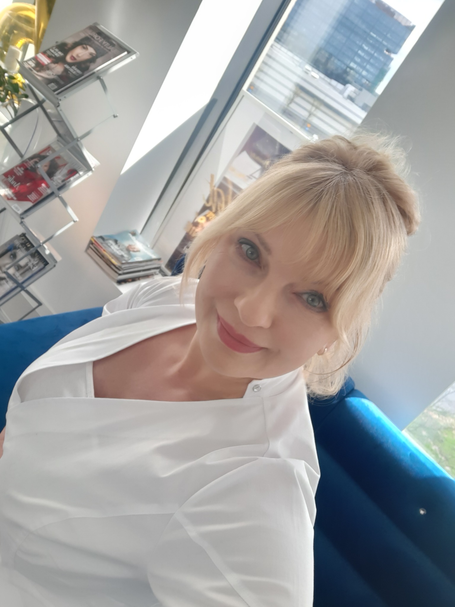 Uśmiechnięta blondynka w białym uniformie, siedząca na niebieskiej sofie w salonie z magazynami i widokiem na miasto.