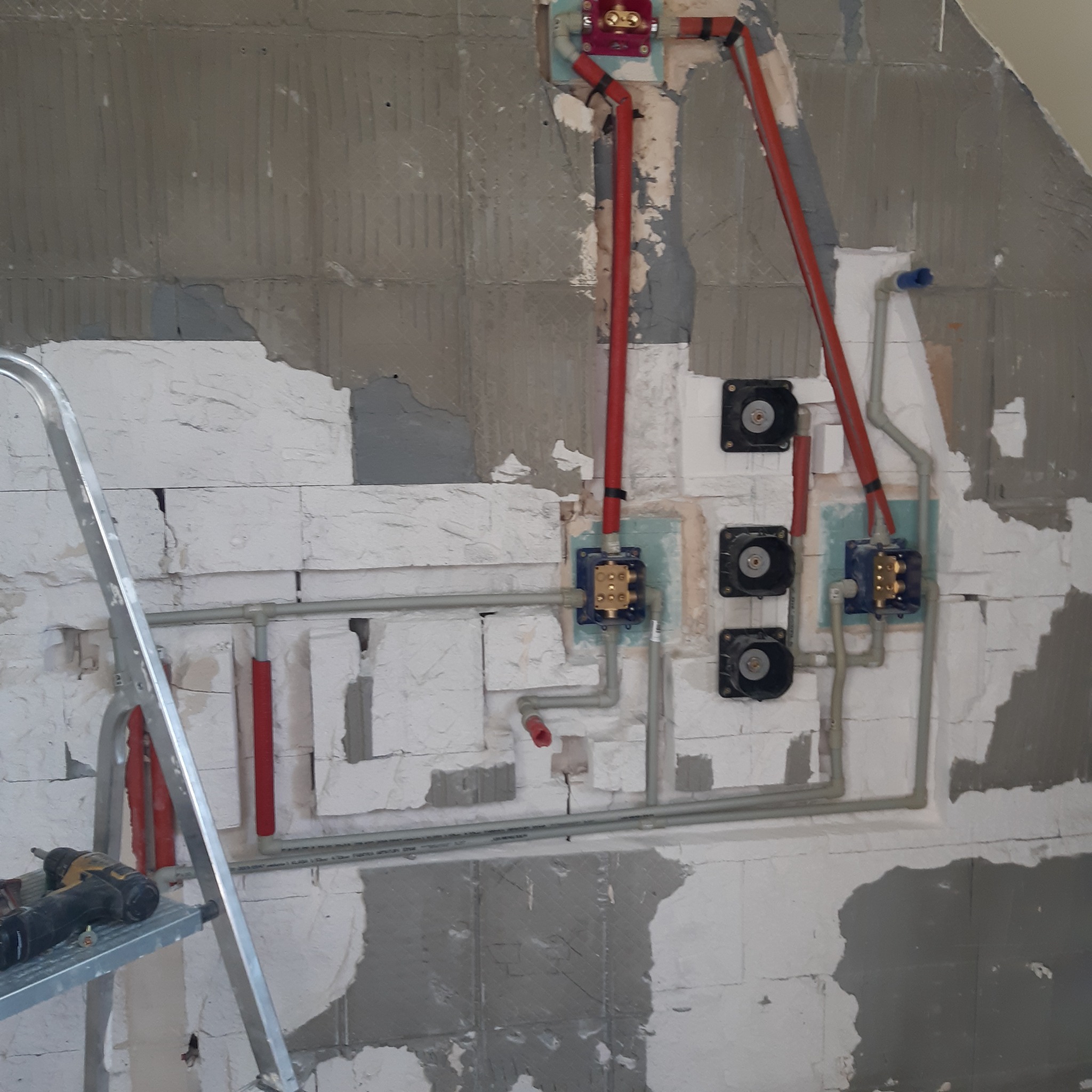 Instalacja wodno-kanalizacyjna w trakcie prac w łazience w Lubinie, widoczne rury, zawory, fragment ściany z niedokończonymi pracami murarskimi oraz drabina z wiertarką.