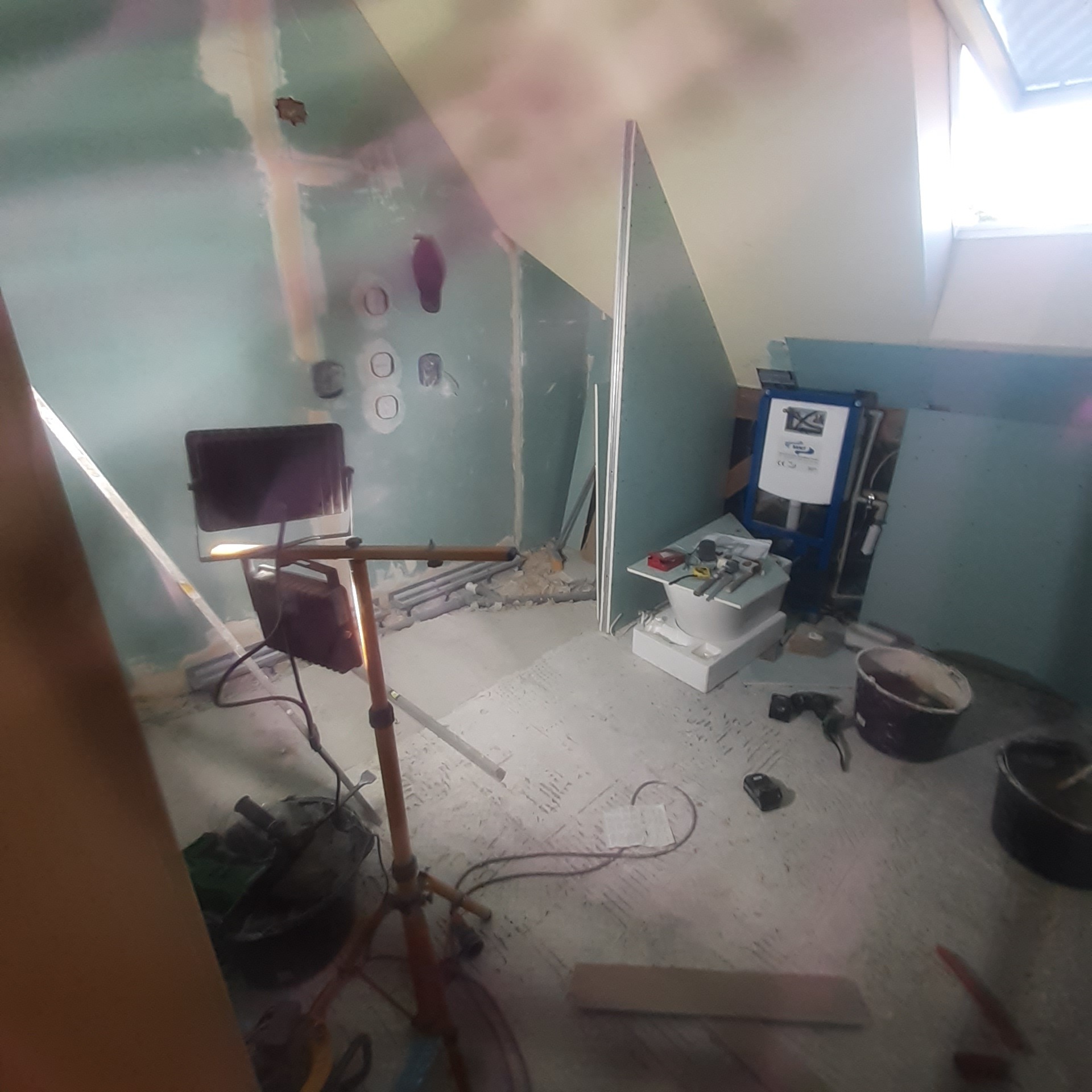 Łazienka w trakcie remontu: widoczne narzędzia, płyty gipsowe, stelaż podtynkowy WC, lampa halogenowa i ślady po instalacji elektrycznej na ścianie.