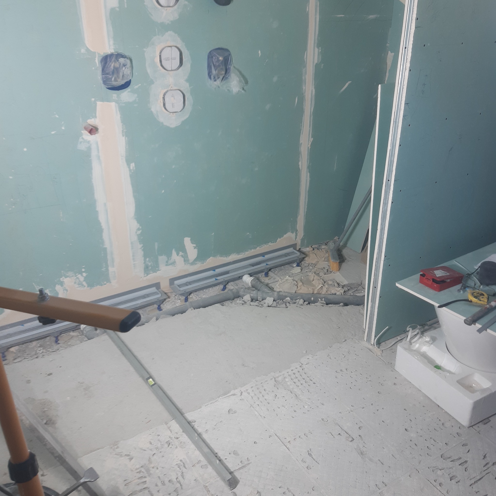 Niedokończona łazienka z widocznymi odpływami liniowymi w podłodze, rurami, płytami gipsowo-kartonowymi na ścianach i narzędziami pracy.