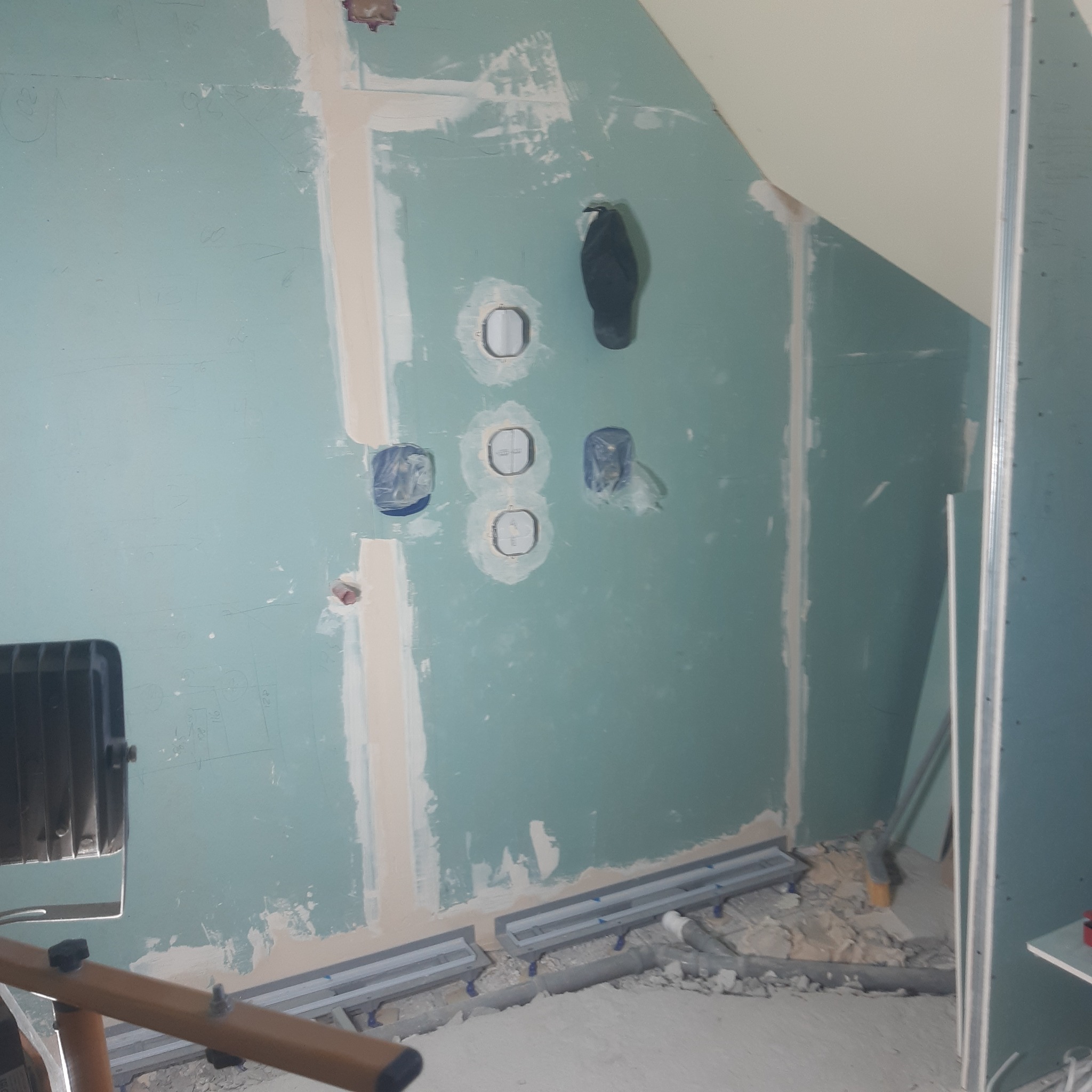 Ściana pokryta niebieską płytą karton-gips z widocznymi puszkami elektrycznymi zabezpieczonymi folią, szpachlowanymi łączeniami i metalowymi profilami podłogowymi w trakcie montażu.