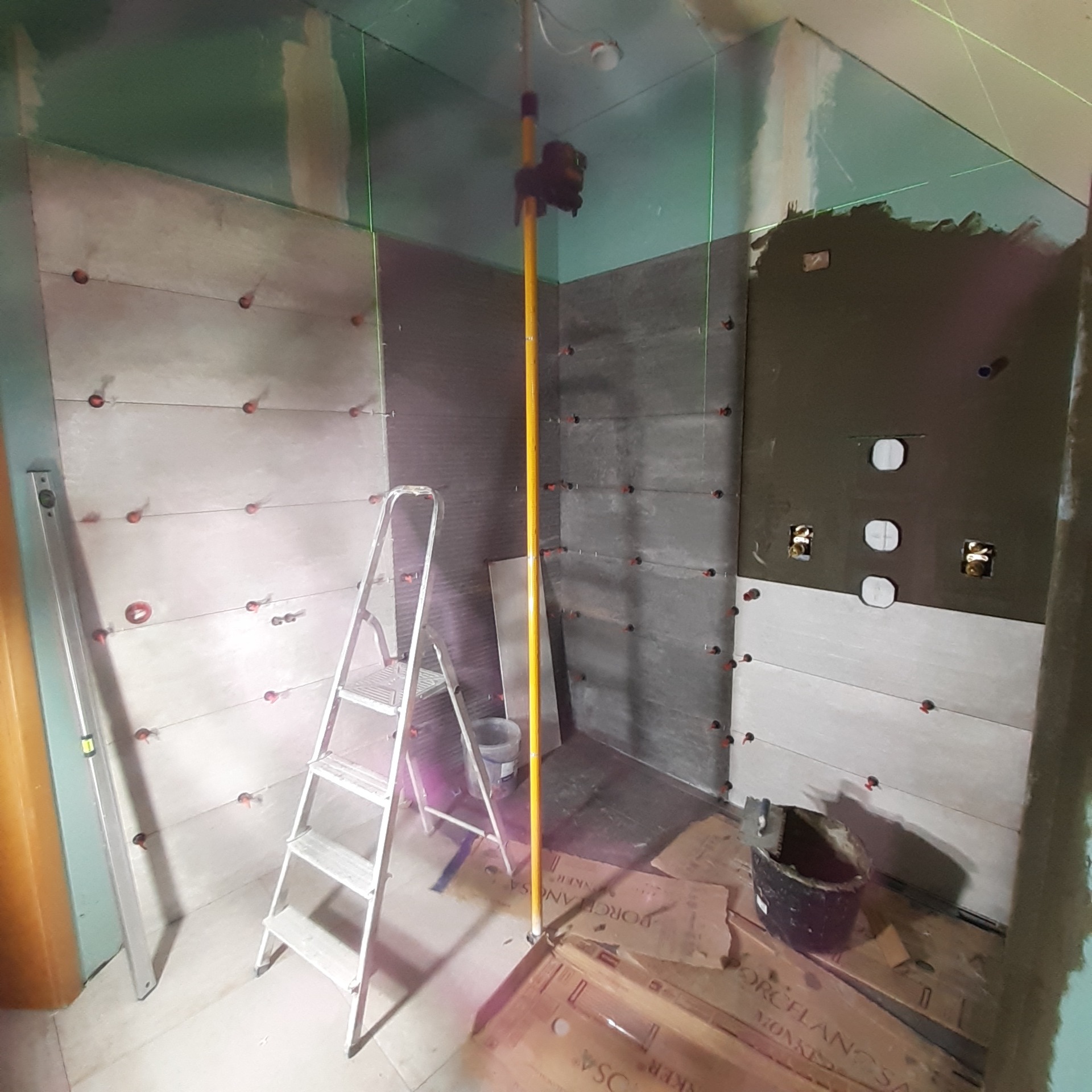 Łazienka w trakcie remontu: układanie płytek imitujących kamień, poziomowanie laserowe, drabina, wiadro z zaprawą, kartony po płytkach.