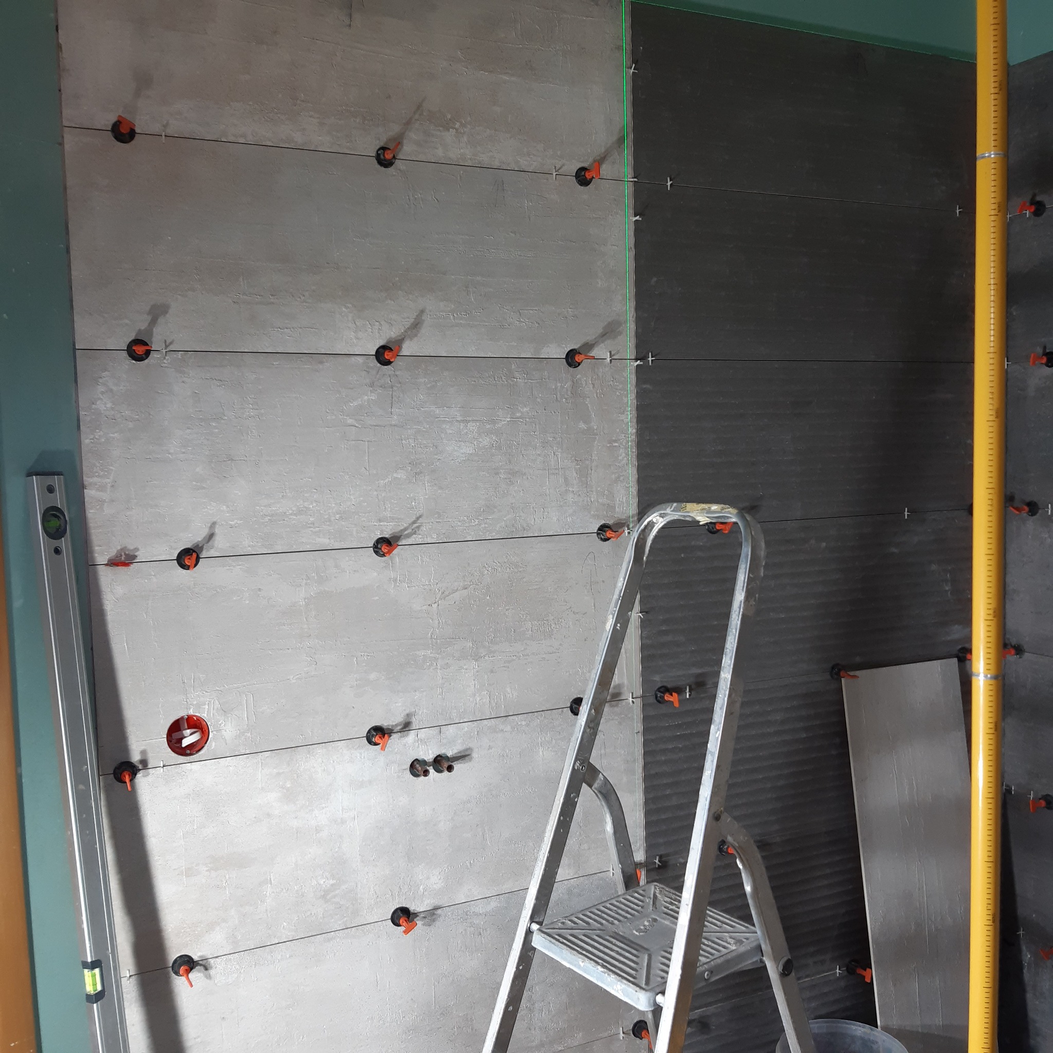 Układanie płytek imitujących drewno na ścianie z użyciem systemu poziomowania, widoczna drabina i poziomica.