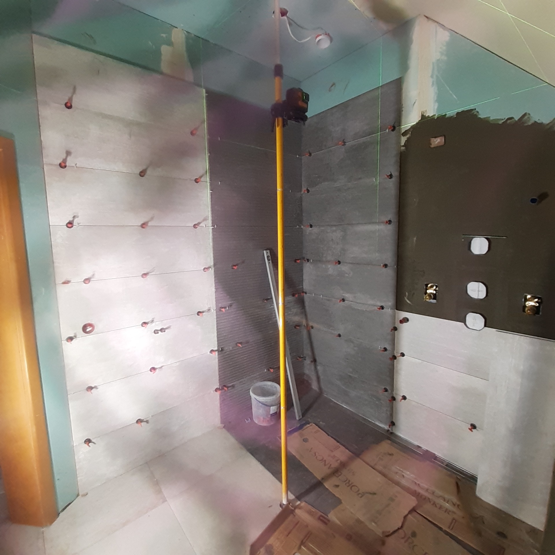 W trakcie układania szarych płytek w łazience z użyciem poziomicy laserowej i krzyżyków dystansowych, widoczne puszki elektryczne i fragment opakowania płytek na podłodze.