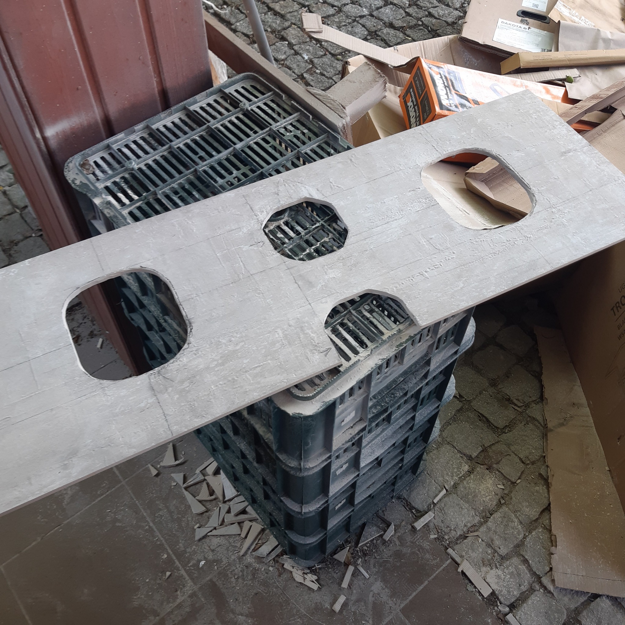 Płyta z wyciętymi otworami, prawdopodobnie blat łazienkowy, leżąca na plastikowych skrzynkach w otoczeniu gruzu i kartonów.