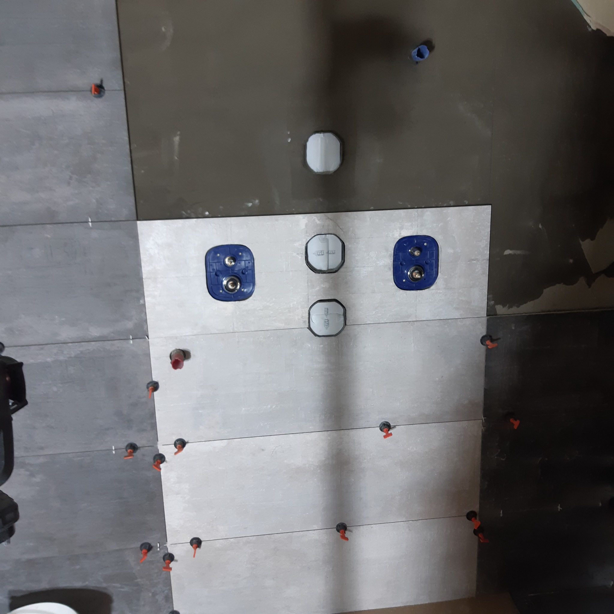 Montaż płytek ceramicznych na ścianie z widocznymi otworami na instalacje, użyte klipsy poziomujące i krzyżyki dystansowe, fragment ściany z ciemniejszymi płytkami.