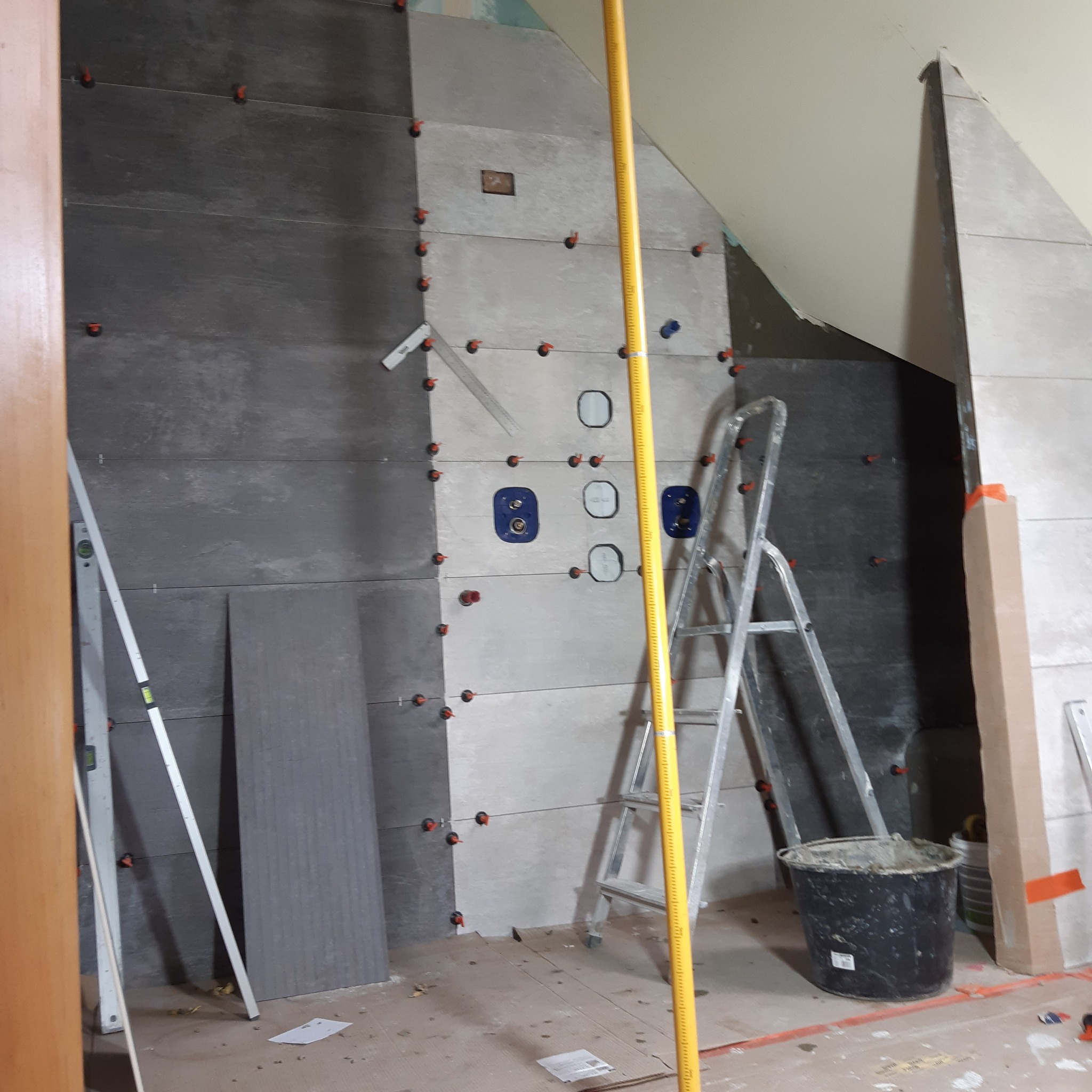 Ściana łazienki w trakcie układania szarych płytek, widoczne poziomice, drabina, wiadro z zaprawą i elementy mocujące płytki, w tle instalacja wodna.