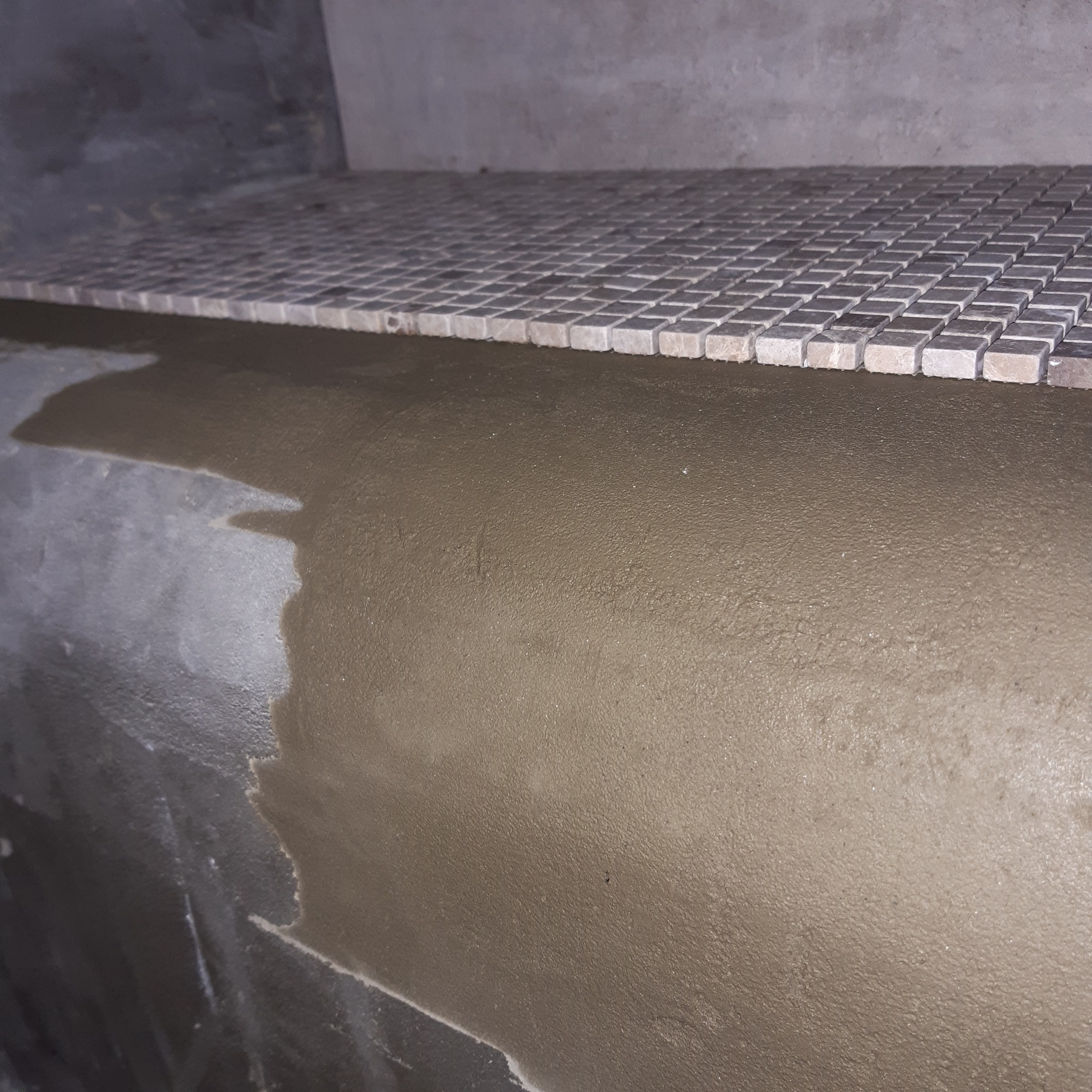 Przygotowanie podłoża pod mozaikę w łazience: warstwa kleju na ścianie, ułożona siatka z mozaiką ceramiczną w odcieniach szarości.