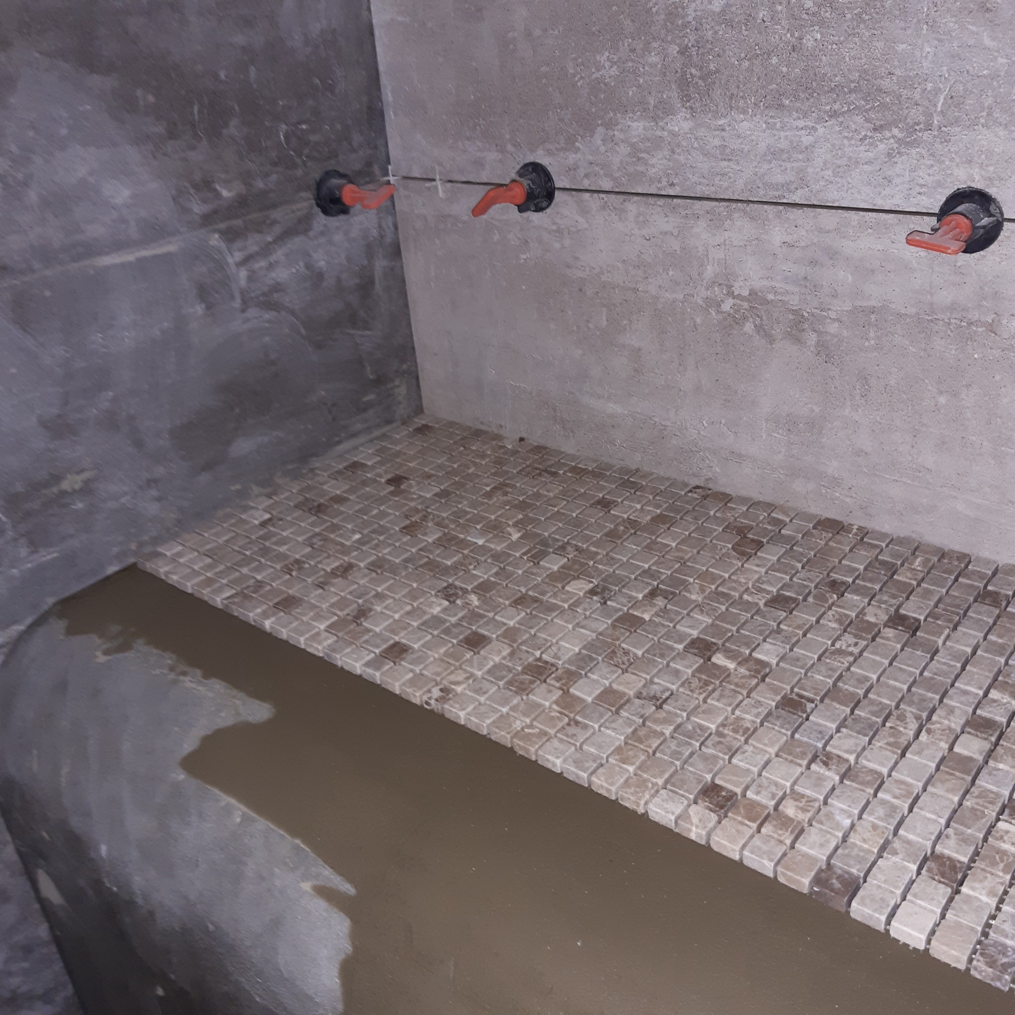 Układanie mozaiki z drobnych płytek w odcieniach beżu i brązu na przygotowanej powierzchni w łazience, widoczne krzyżyki dystansowe i wystające rury instalacji wodnej.