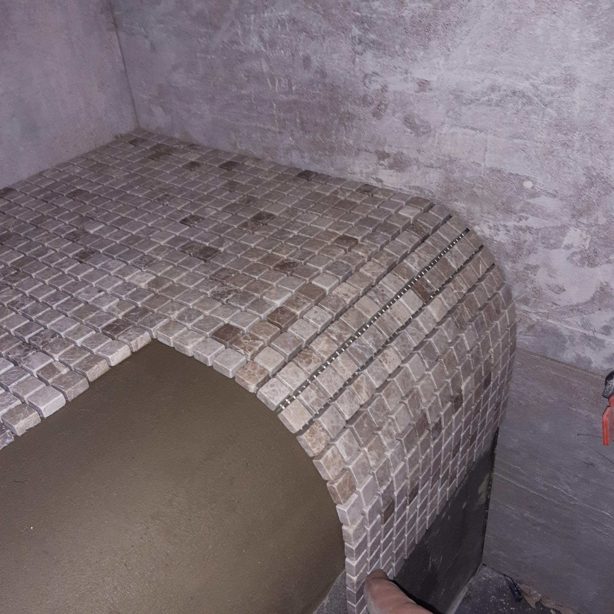 Mozaika z jasnych, kwadratowych płytek ułożona na zaokrąglonej powierzchni w rogu łazienki, widoczny fragment niezabezpieczonej ściany i fugi pomiędzy płytkami.