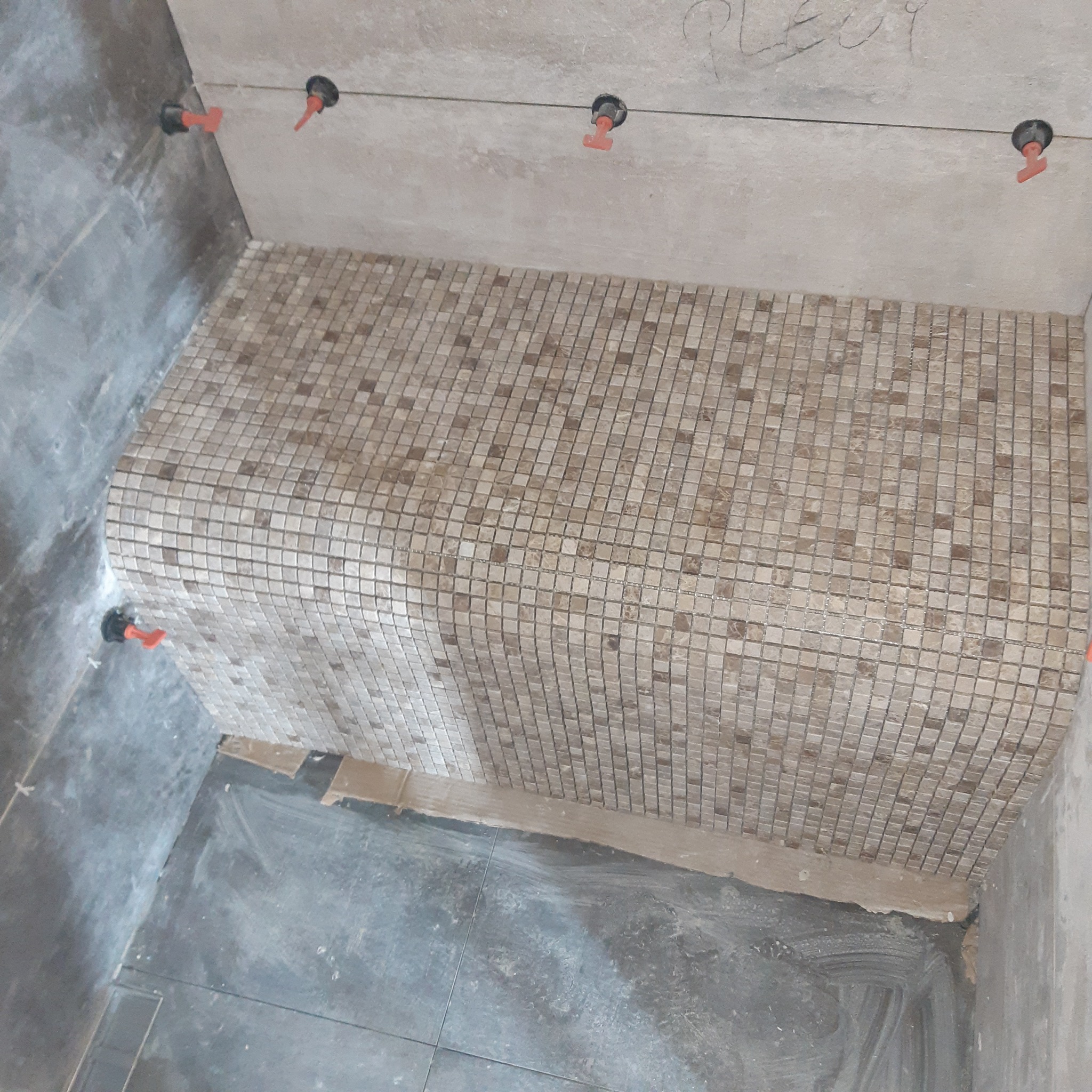 Mozaika z drobnych, kwadratowych płytek w odcieniach beżu i brązu, ułożona na zaokrąglonej krawędzi w łazience, z widocznymi systemami poziomowania płytek w narożnikach.