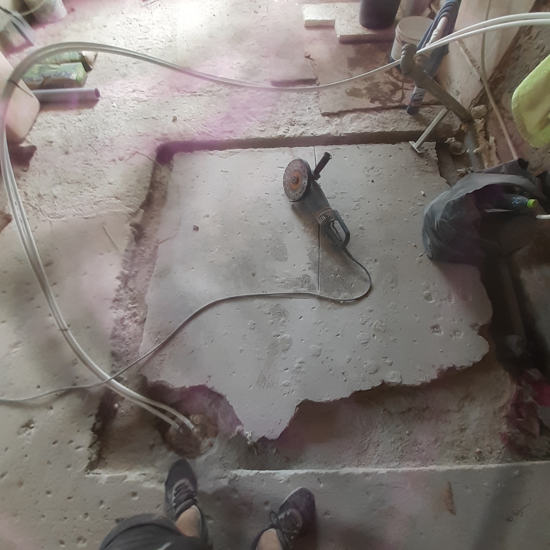 Nierówna, wycięta posadzka betonowa z widoczną szlifierką kątową, fragmentami instalacji oraz obuwiem roboczym w rogu kadru.