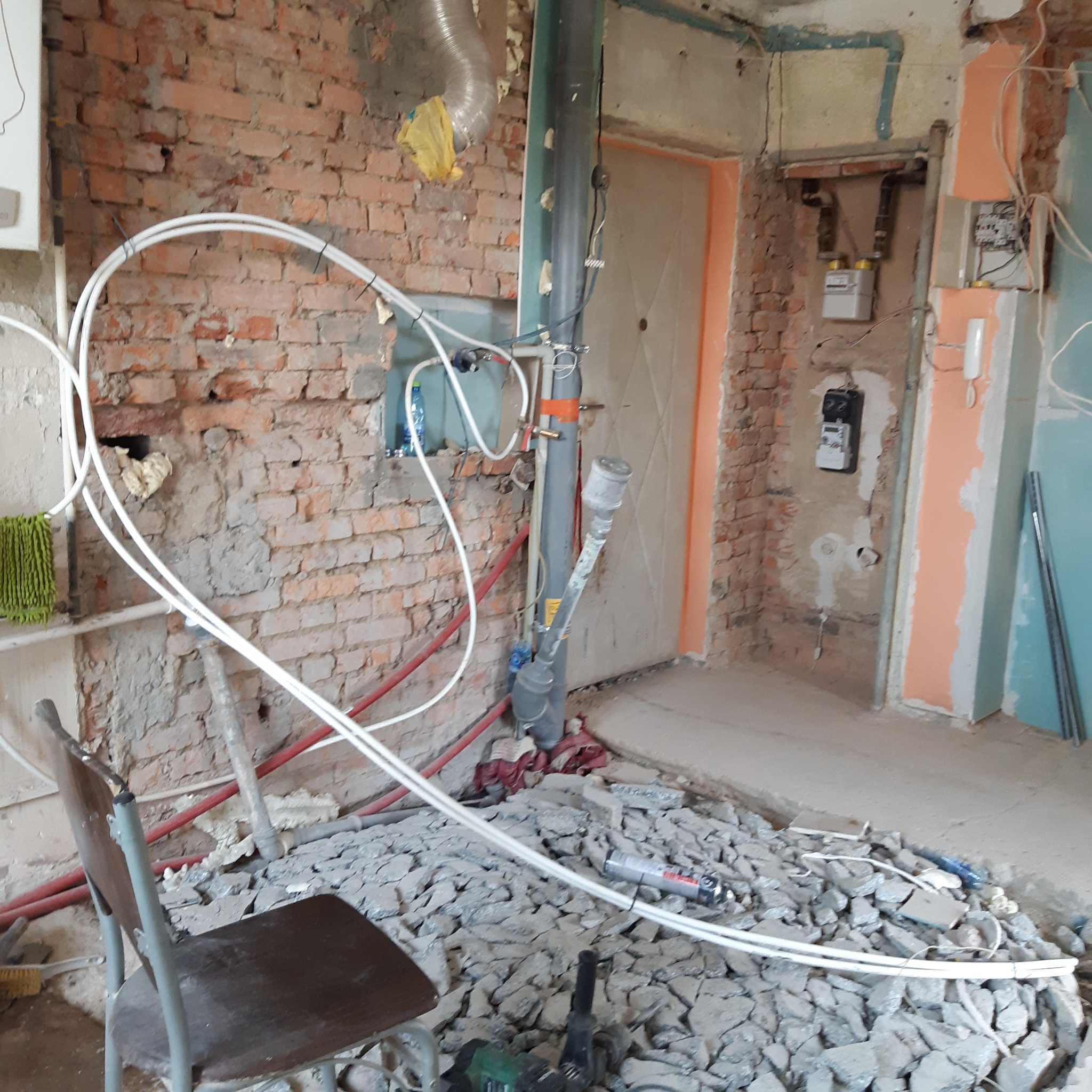 Wnętrze w trakcie demontażu: odsłonięta cegła, rury, gruz na podłodze, krzesło i narzędzia.