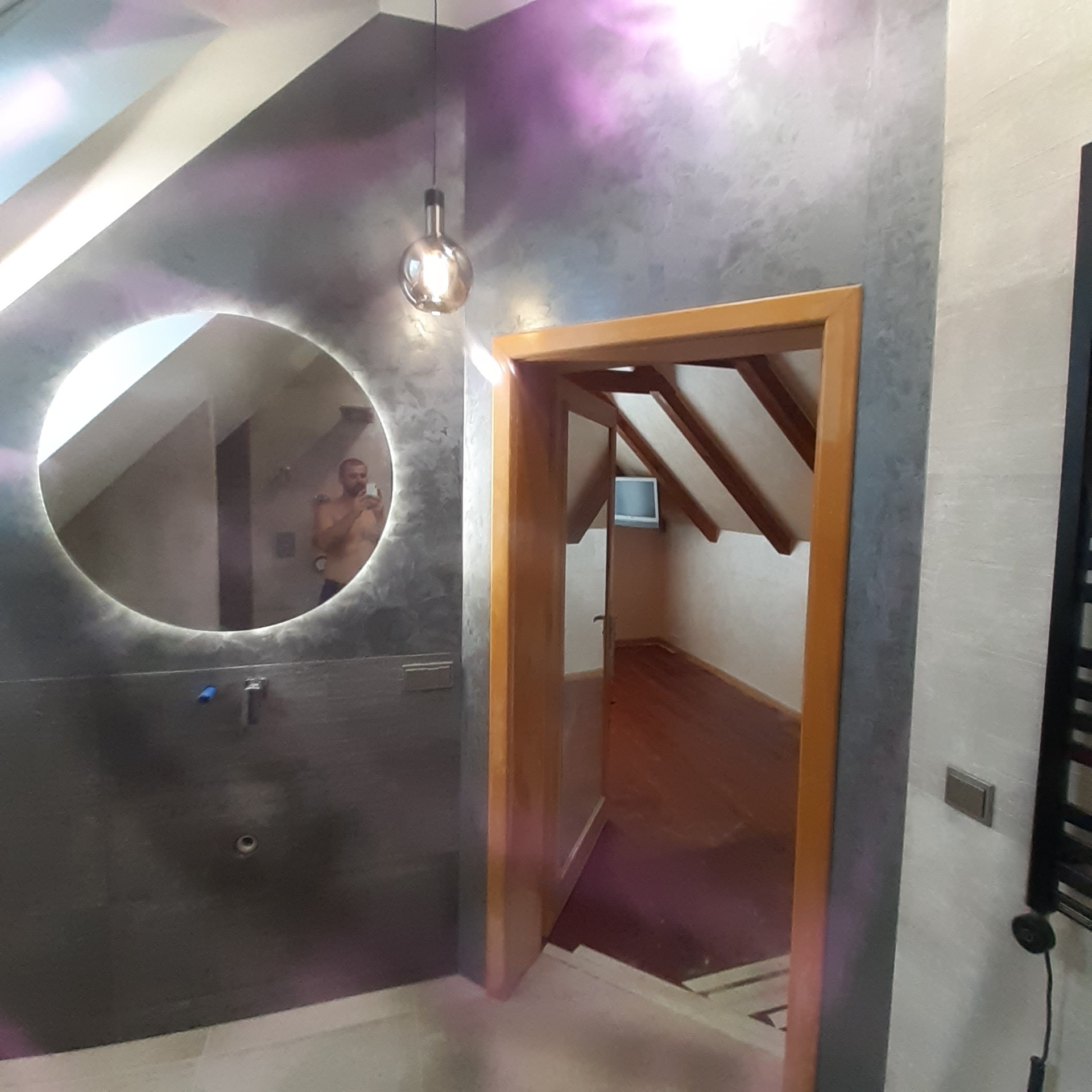 Nowoczesna łazienka na poddaszu z okrągłym lustrem z podświetleniem LED, szarą ścianą z efektem dekoracyjnym i widokiem na pokój przez drewniane drzwi.