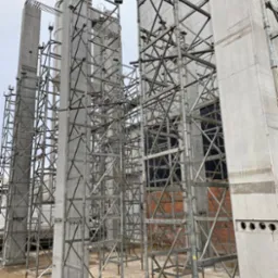 Surowe betonowe słupy konstrukcyjne w trakcie budowy, otoczone metalowymi rusztowaniami na placu budowy, widoczne elementy ceglanej ściany w tle.