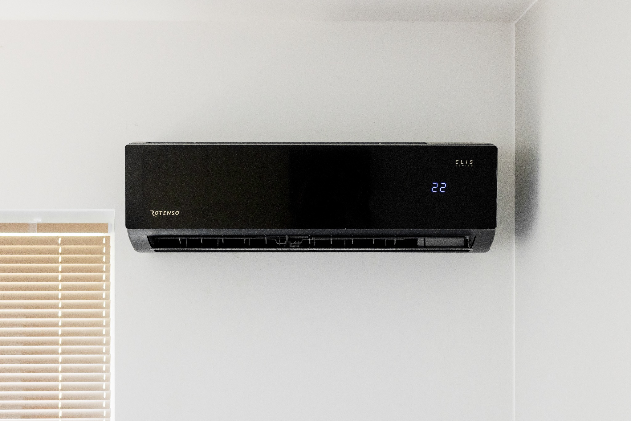 Czarny klimatyzator Rotenso z wyświetlaczem temperatury 22 stopnie, zamontowany na białej ścianie, obok okna z żaluzjami.