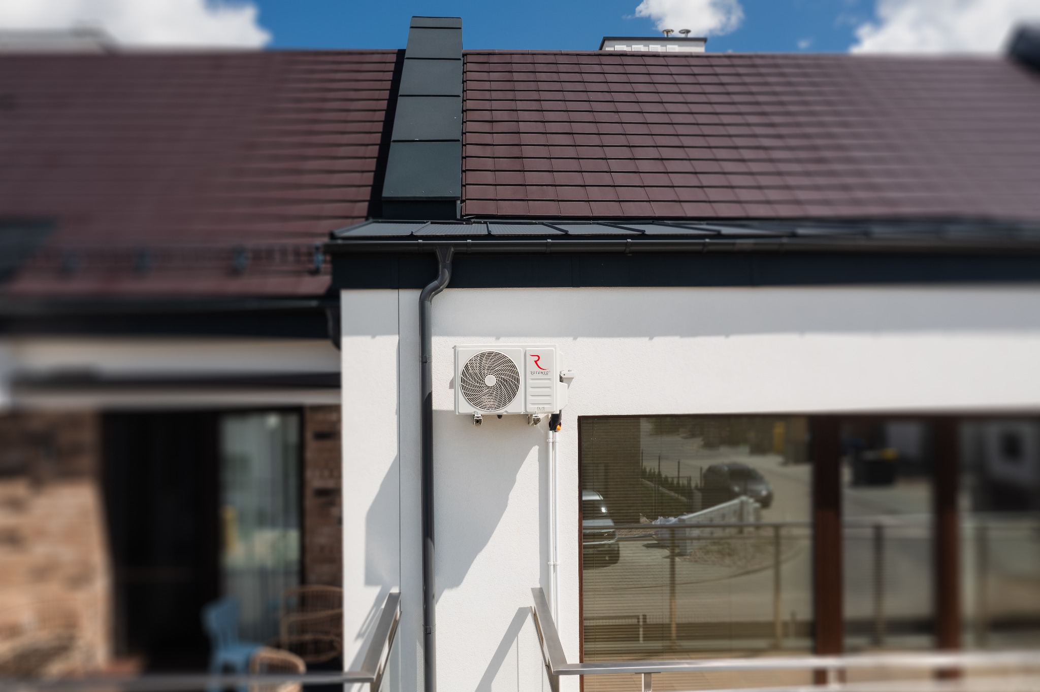 Zewnętrzna jednostka klimatyzacji Rotenso zamontowana na białej ścianie domu z brązowym dachem, widok z balkonu.